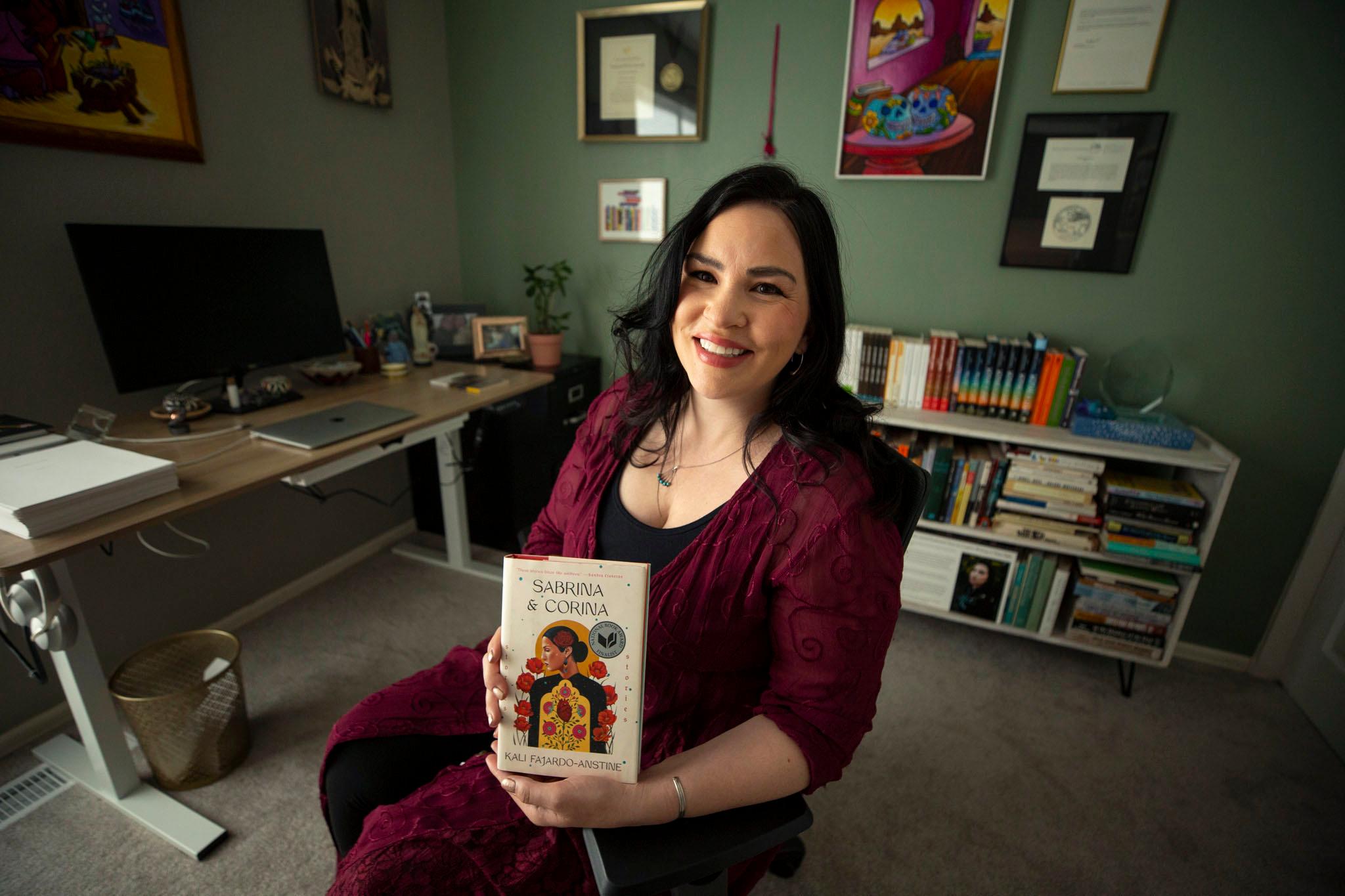 A woman with dark hair and dark eyes looks right at the camera and holds a book as she sits in a green painted room.