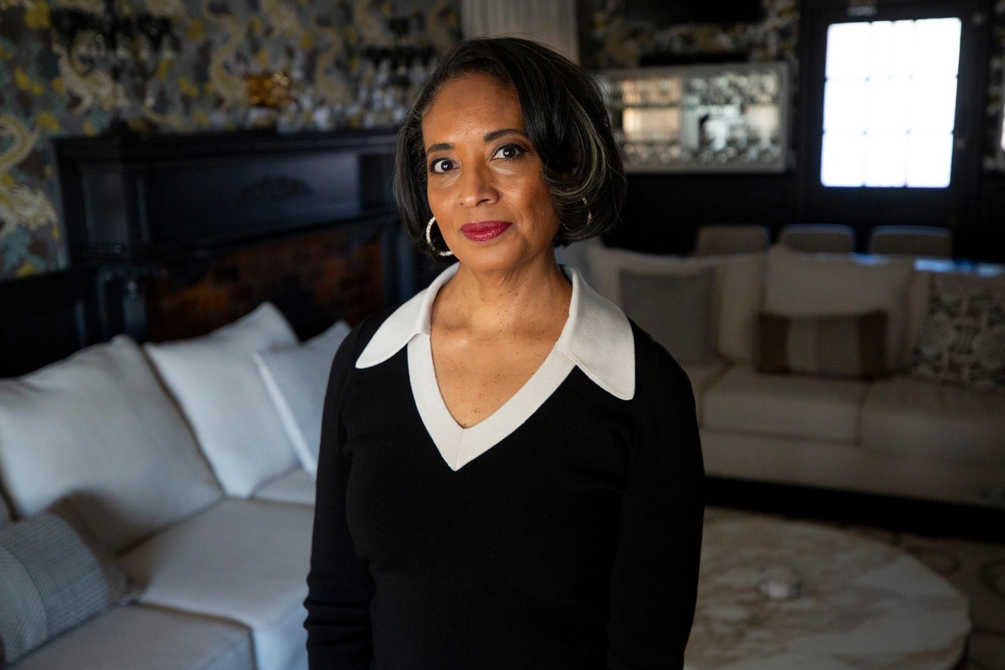 A woman in a black outfit with a large white collar stands in an opulent room of wood and patterned wallpaper as she looks into the camera.
