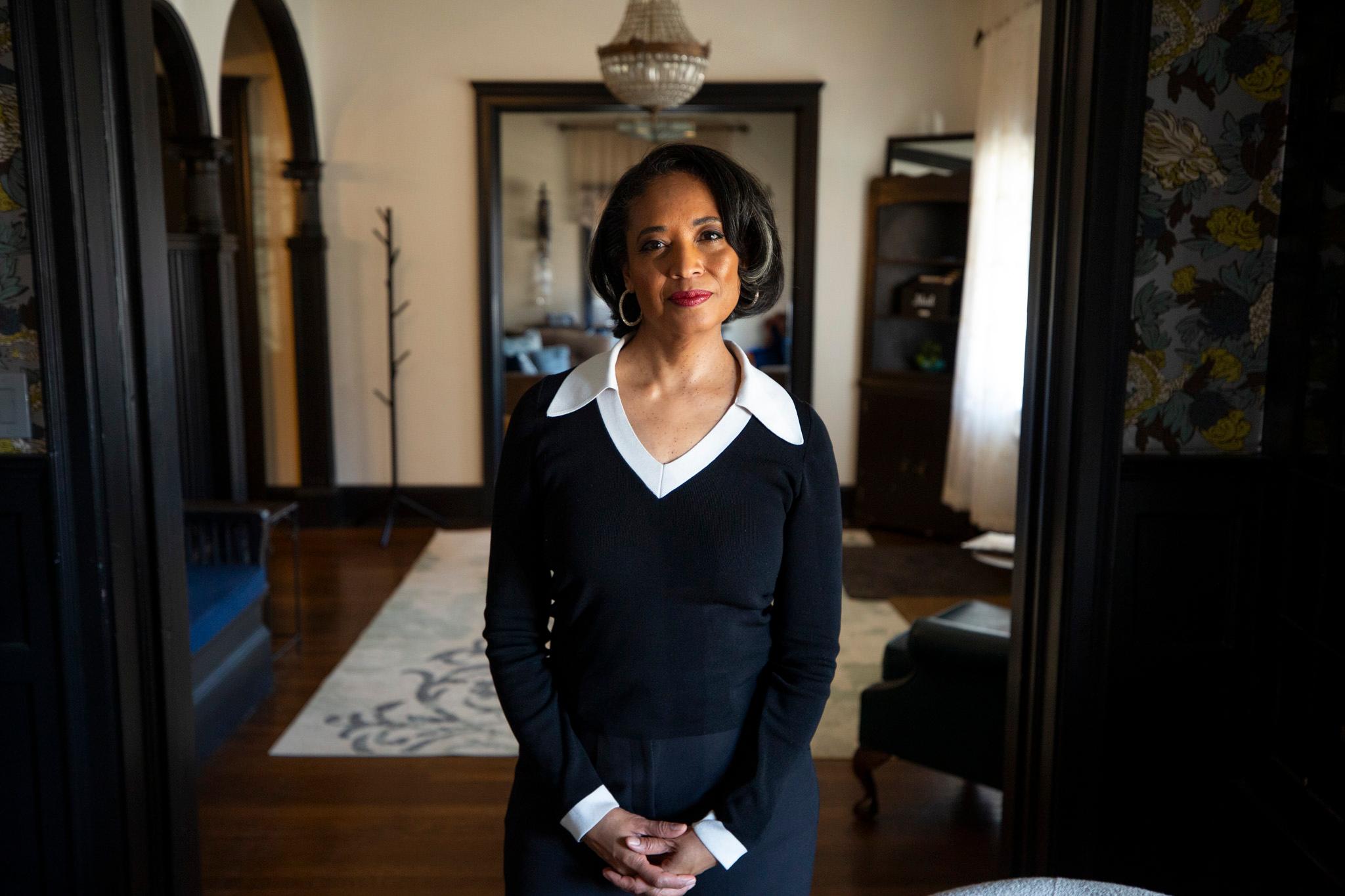 A woman in a black outfit with a large white collar stands in an opulent room of wood and patterned wallpaper as she looks into the camera.