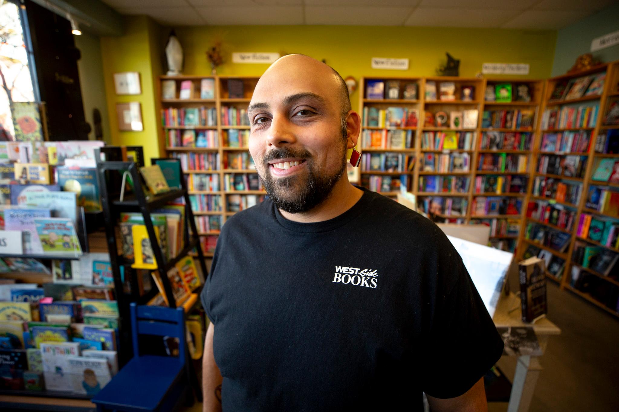 A man in a black shirt that reads "west side books" stands in a warmly lit room filled with bookshelves.