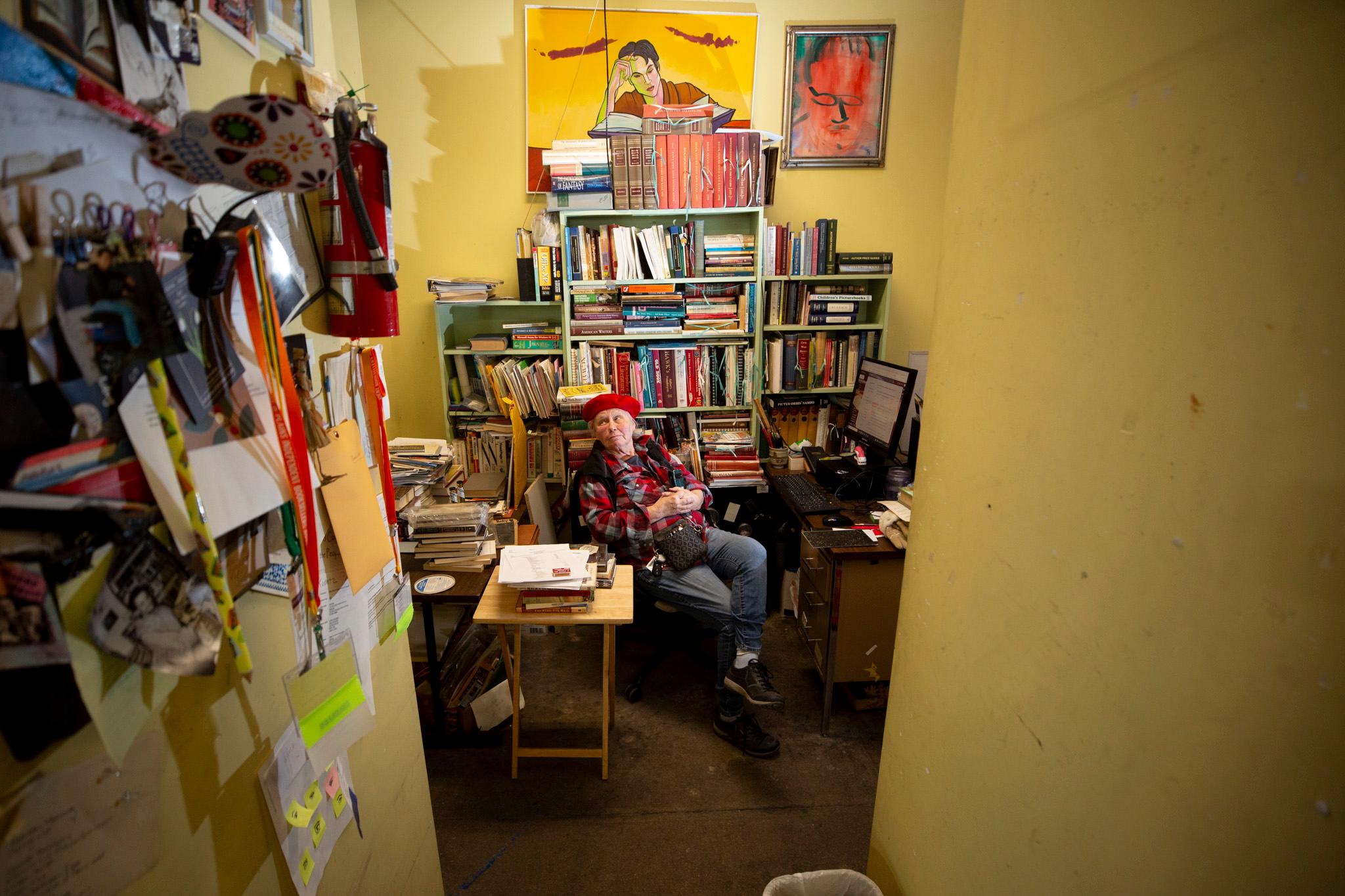 A woman in a red beret sits in a small room down a yellow hallway, surrounded by shelves and stacks of books and art.