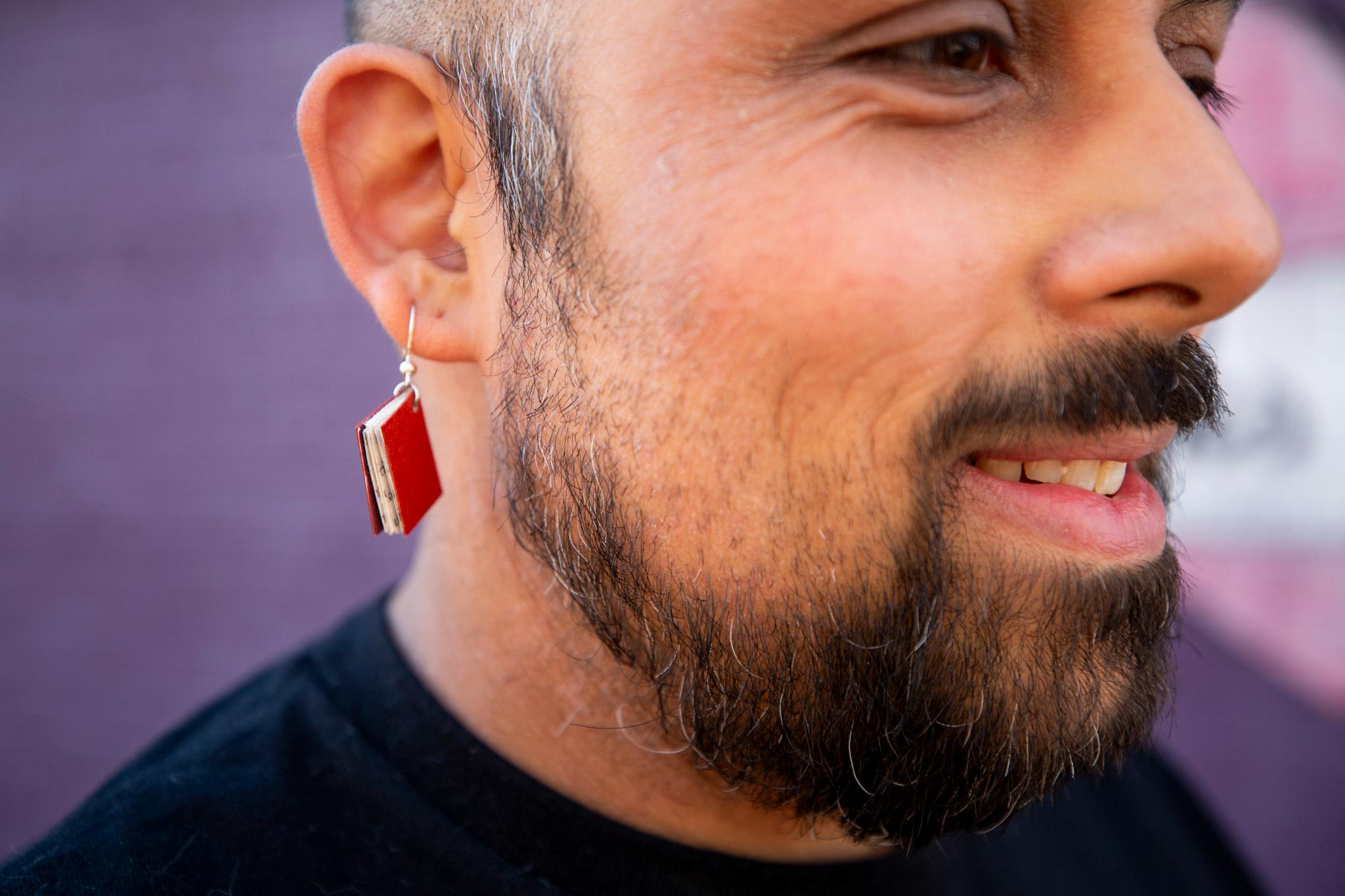A close up of a bearded man, who has a tiny red book earring hanging from his lobe.