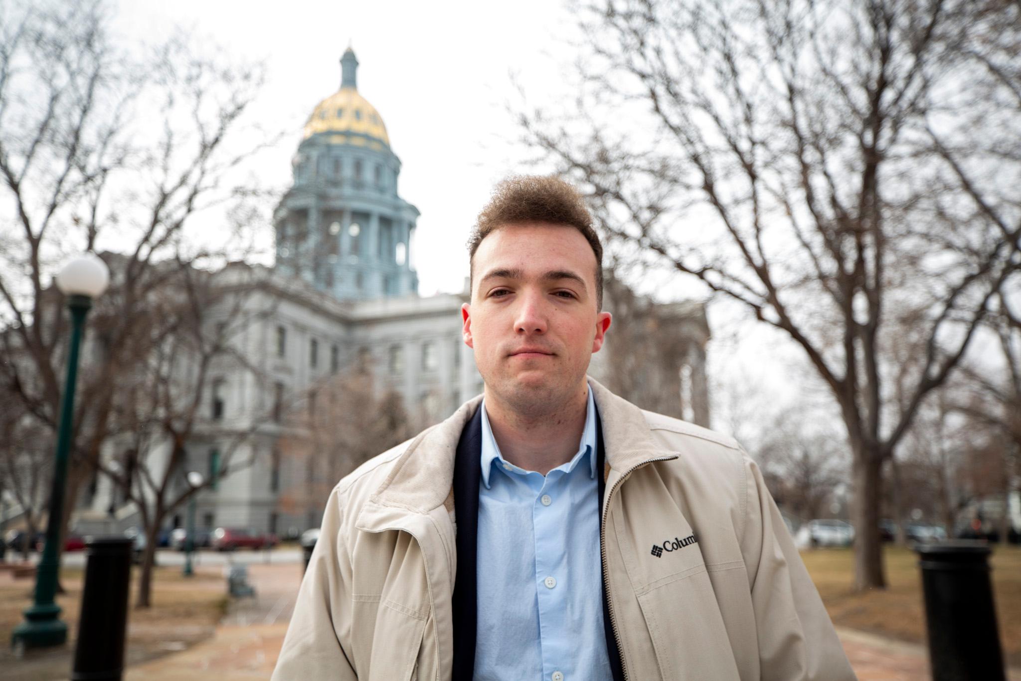 A man in a beige jacket, with a wispy puff of hair on top of his head, stands in front of a large gold-domed building.