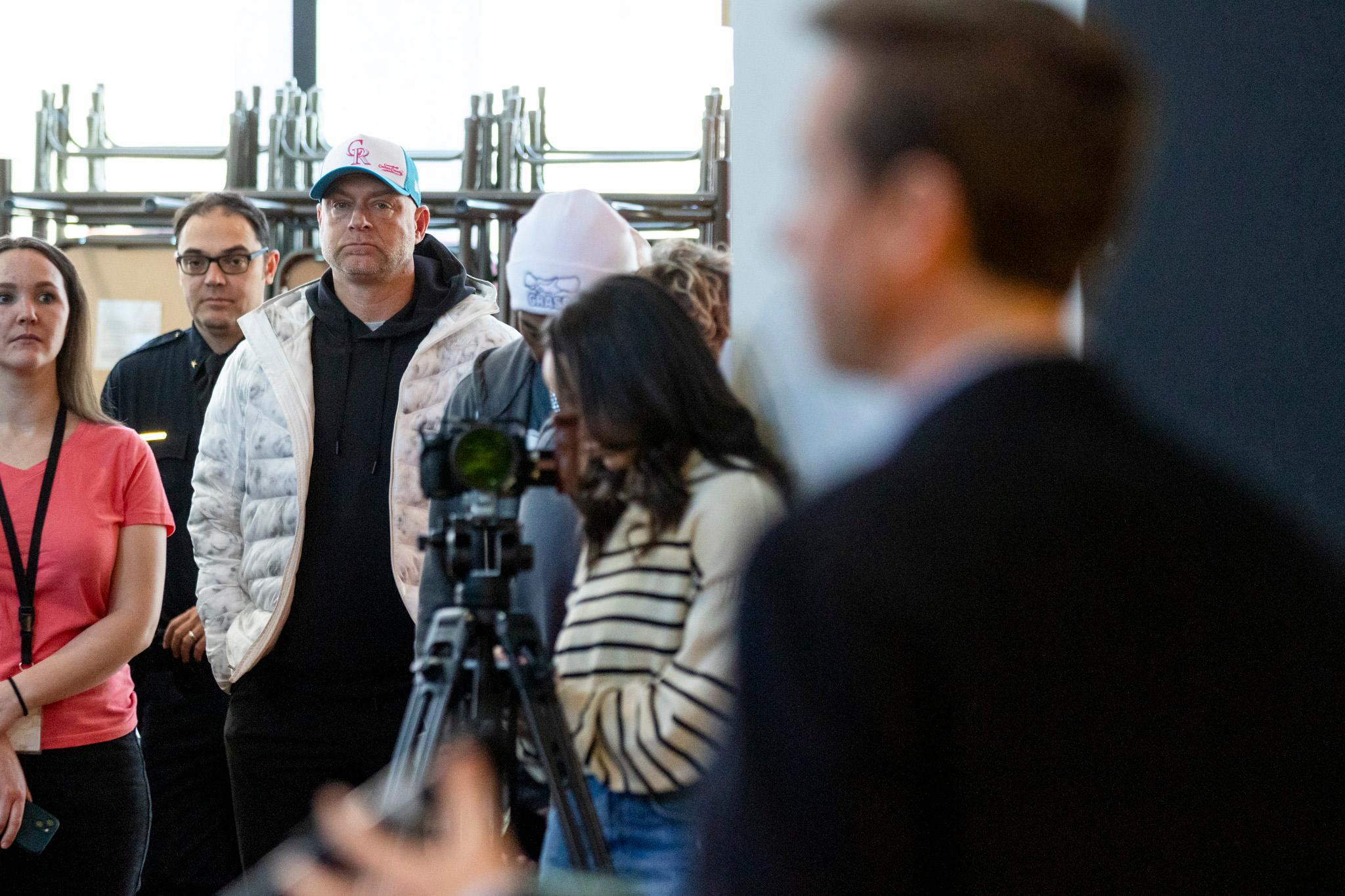 A man in a ballcap watches from afar as another man speaks in the foreground, out of focus.