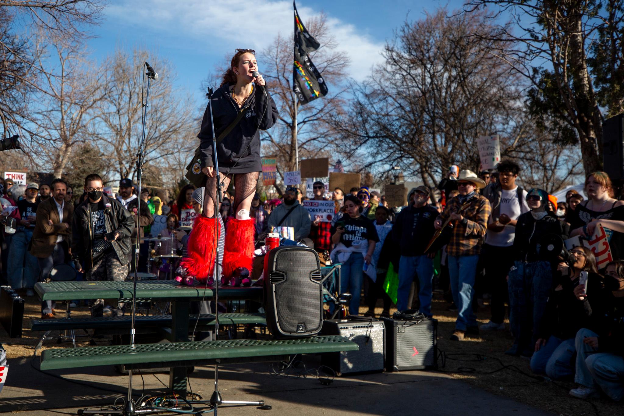A woman in bright red, fuzzy boots yells into a microphone from atop a green picnic table. She's surrounded by a crowd.