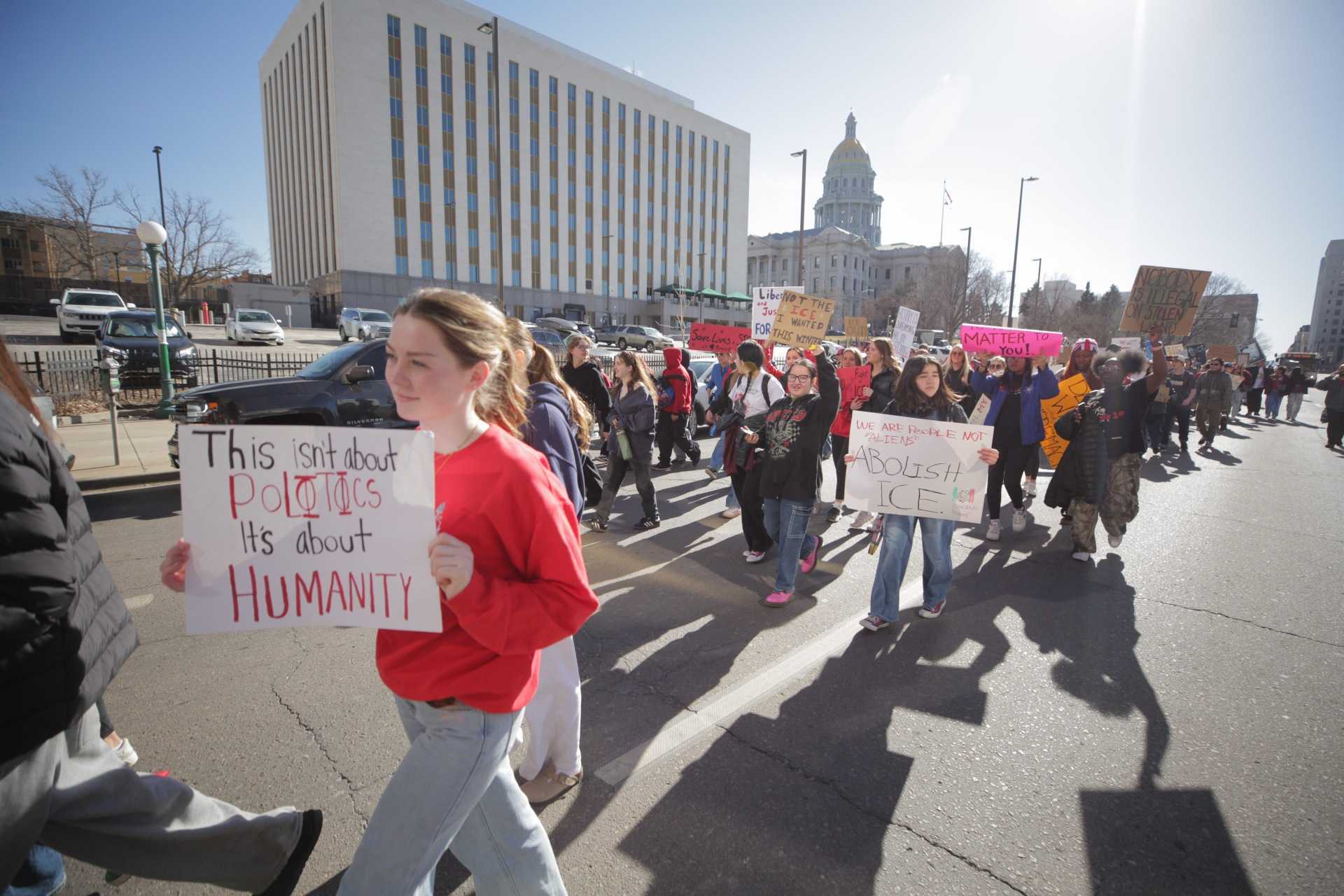 A large crowd of students walk in the street. Many wear red shirts or other articles of clothing. Many hold protest signs. A sign close to the camera reads: "This isn't about politics it's about humanity."