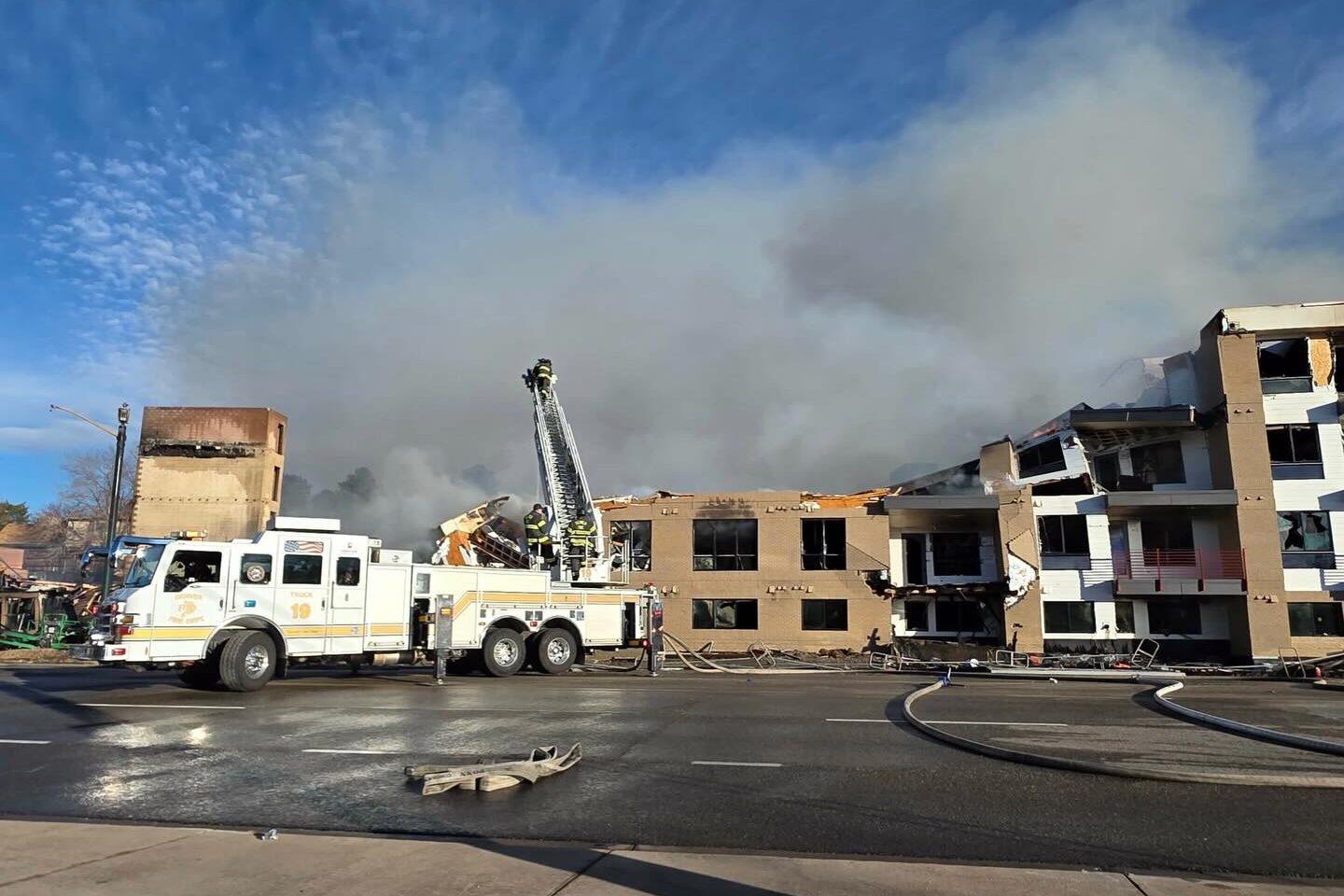 A white fire truck is parked on the side of a road, with a fireman standing on a ladder. The fire truck is connected to a hose, and there is a building under construction nearby.