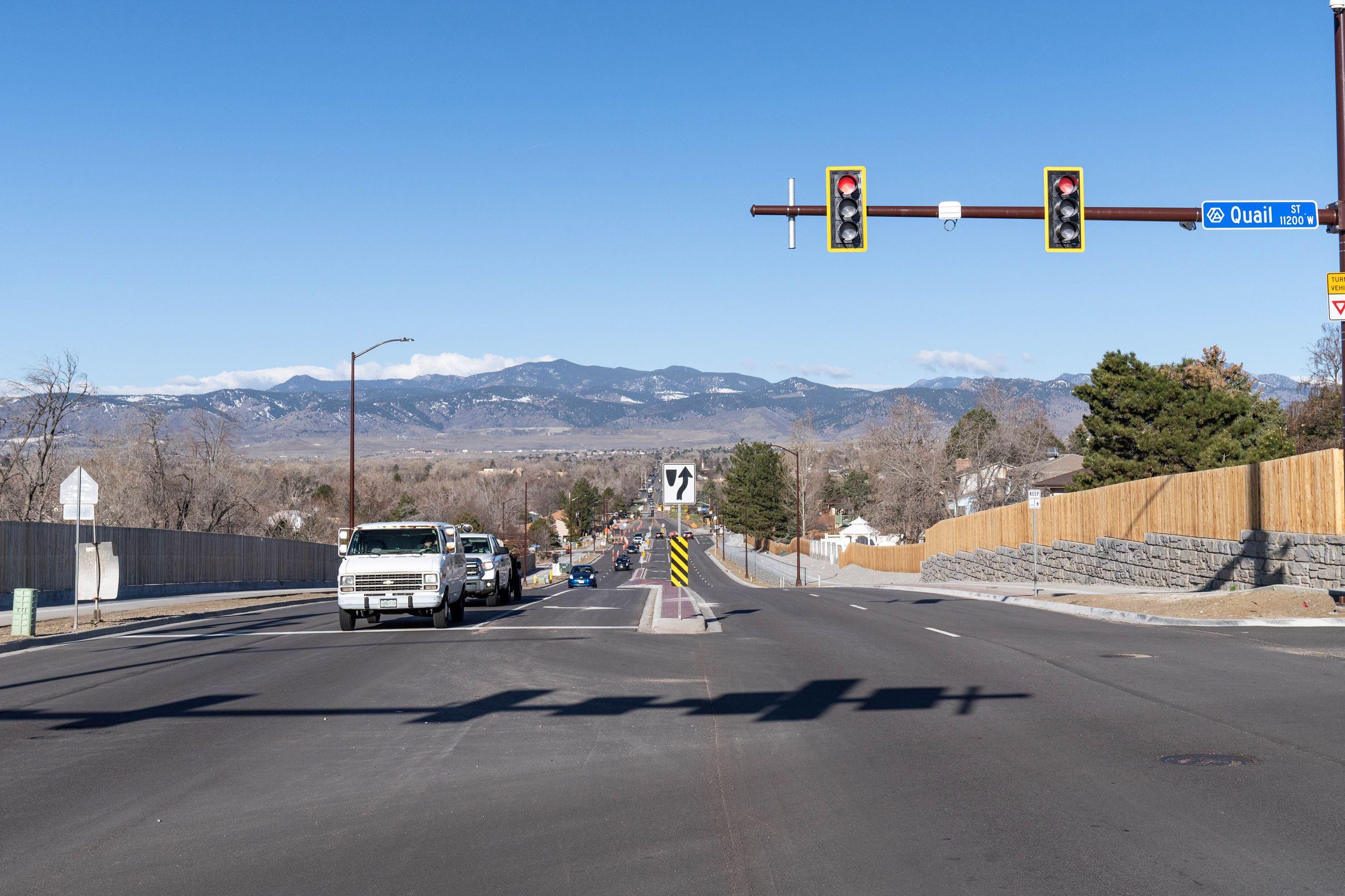 A view of West 72nd Avenue taken from an intersection. The road stretches down the middle of the photo toward a mountain range. Cars line up at the red light on the left side of the image.