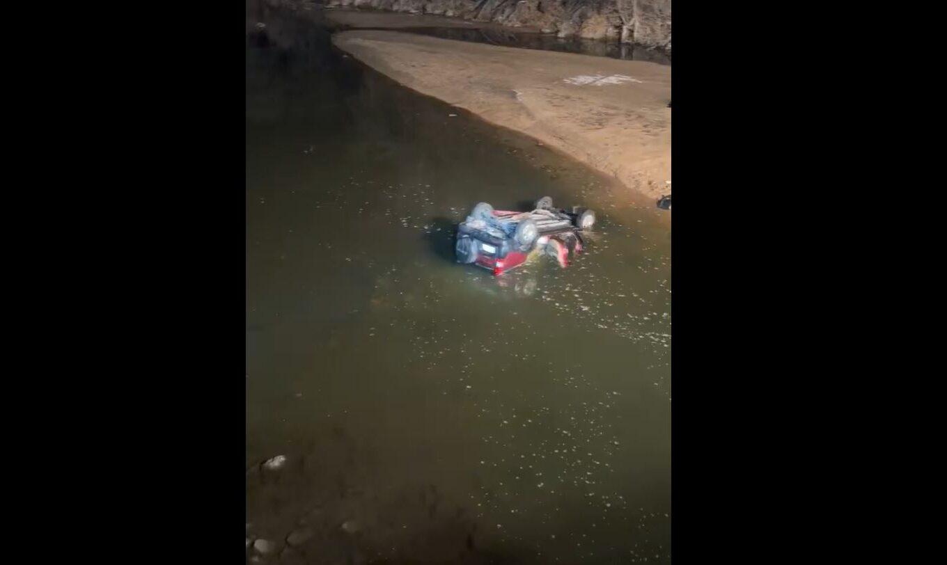 A car, flipped upside down, is halfway submerged into murky water. The wheels are above the water, as well as some of the red car body.