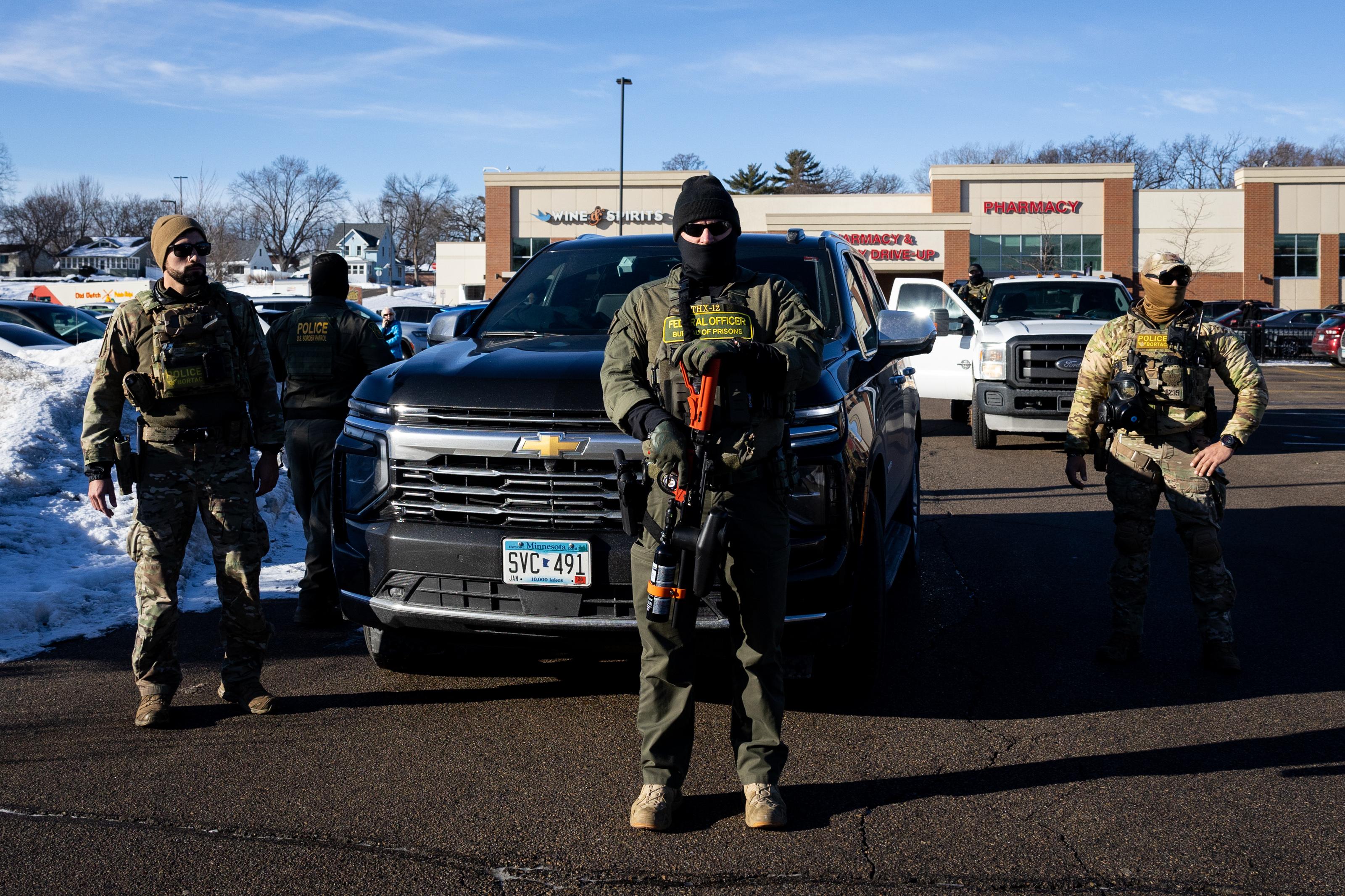 Men in facemasks and tactical vests stand in a parking lot under a blue sky. One looking at the camera holds an air gun, pointed at the ground.