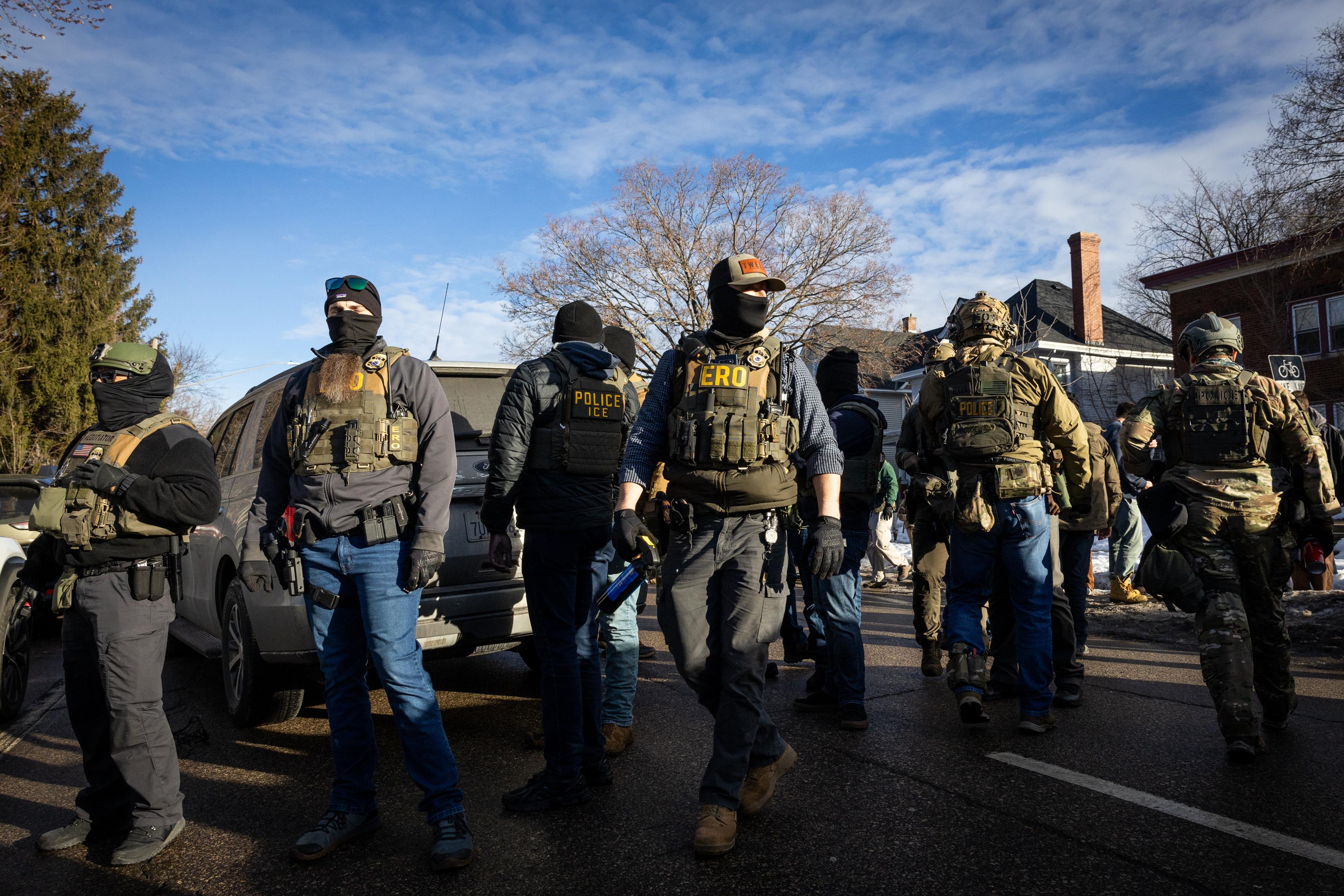 Men in facemasks and tactical vests stand on a street under a blue sky.
