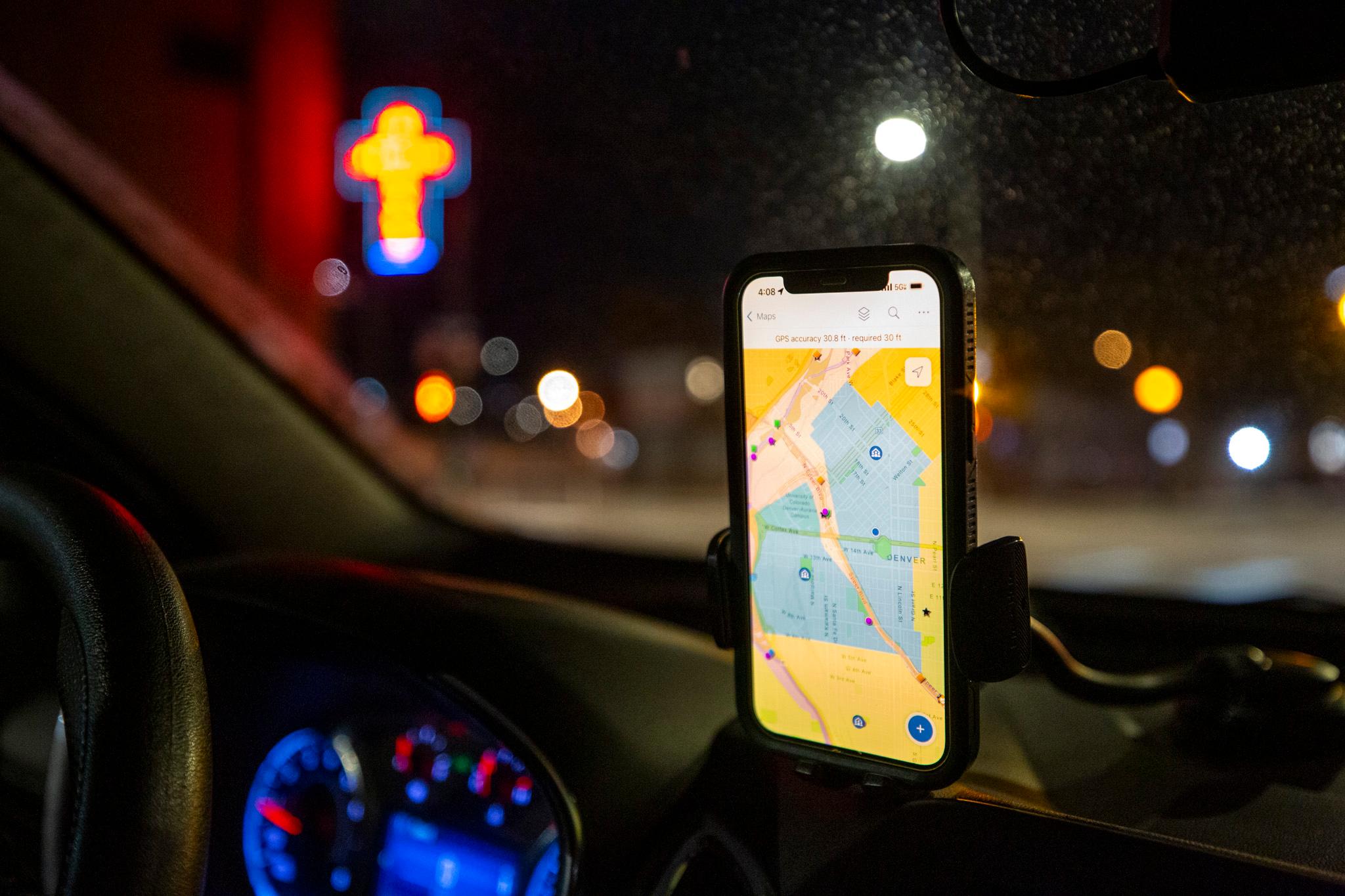 A phone is mounted to a dashboard inside a car. Out of focus, in the background through the window, a neon cross glows in the morning dark.