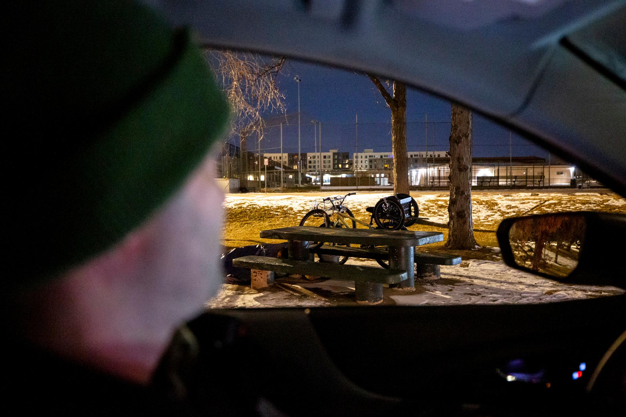 A man in the foreground looks out of a car's window at a picnic bench in a park, with a bike and bike parts leaned up against it. The sky outside is dark.