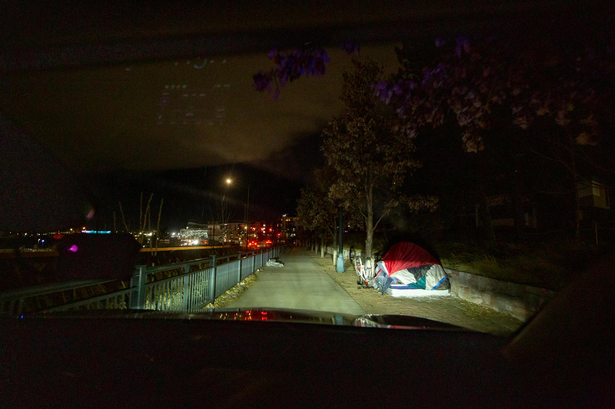 Tent can be seen next to a sidewalk, seen out of a car's windshield. The sky above it is dark.