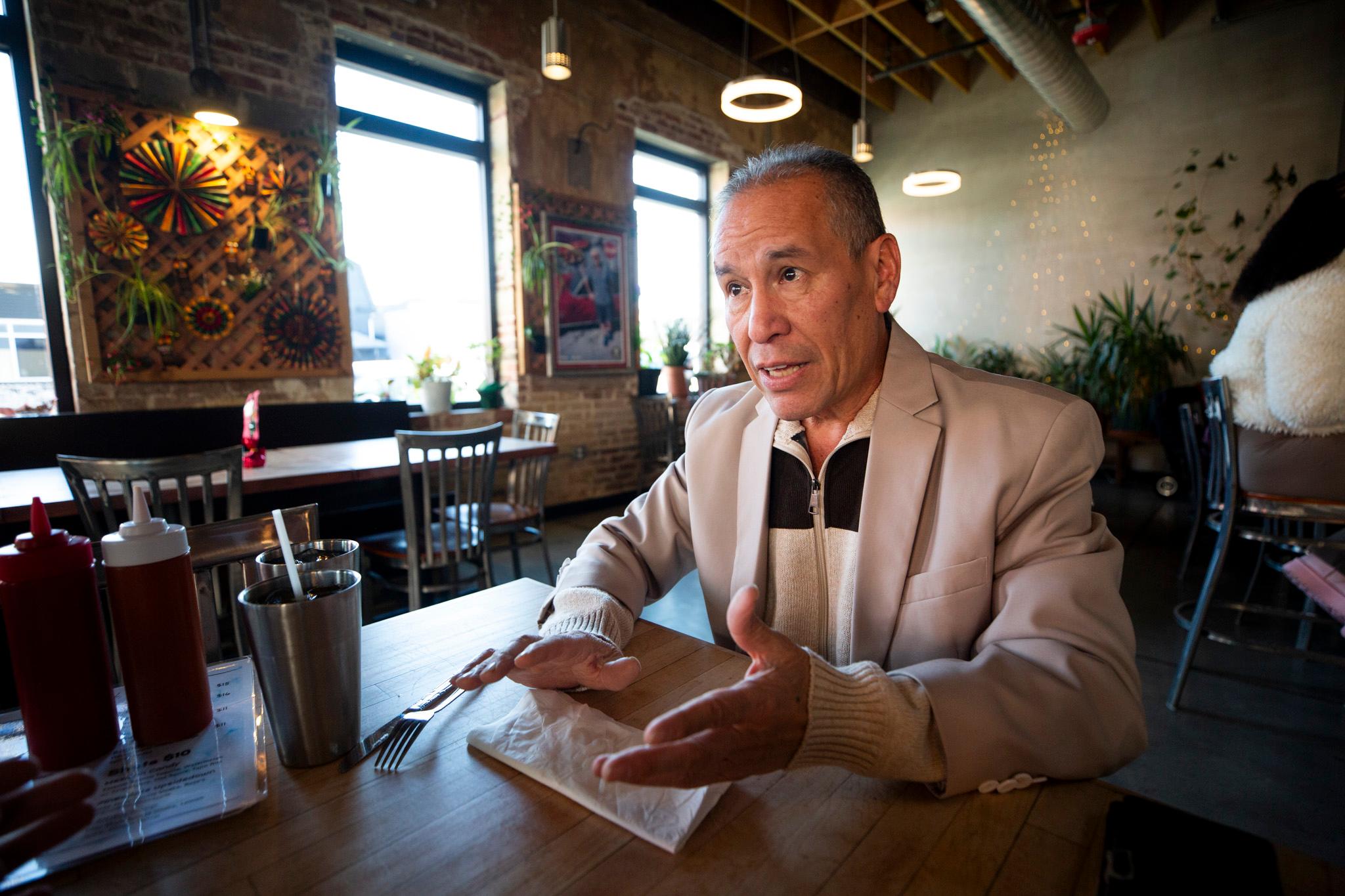 A man in a beige suitjacket sits at a table in a warmly lit room.