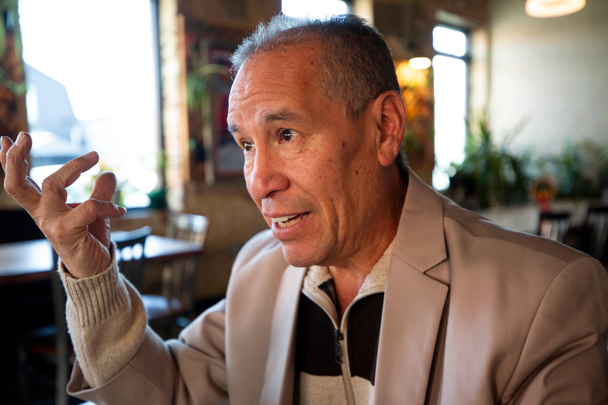 A man in a beige suitjacket sits at a table in a warmly lit room.