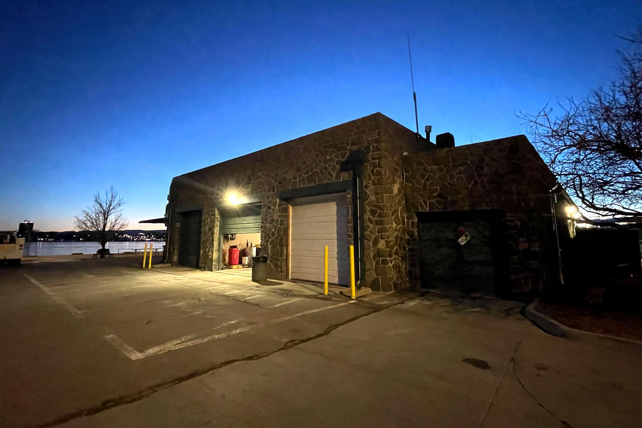 A stone building sits under a fading evening sky. Light pours into the dusk from an open garage door. A lake can be seen in the background.