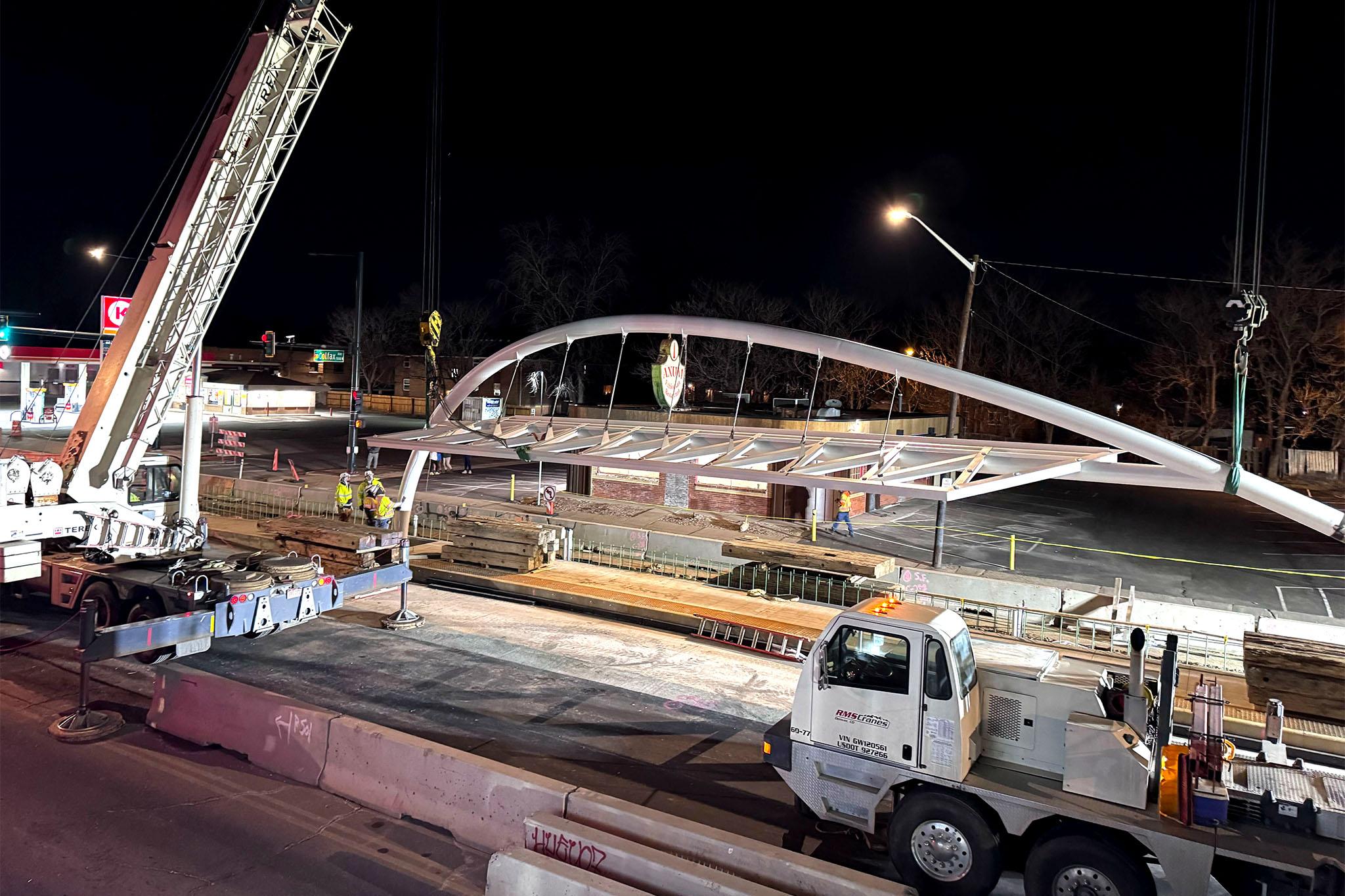 A giant curving piece of metal sits over construction material on a closed asphalt street. The sky above is pitch black.