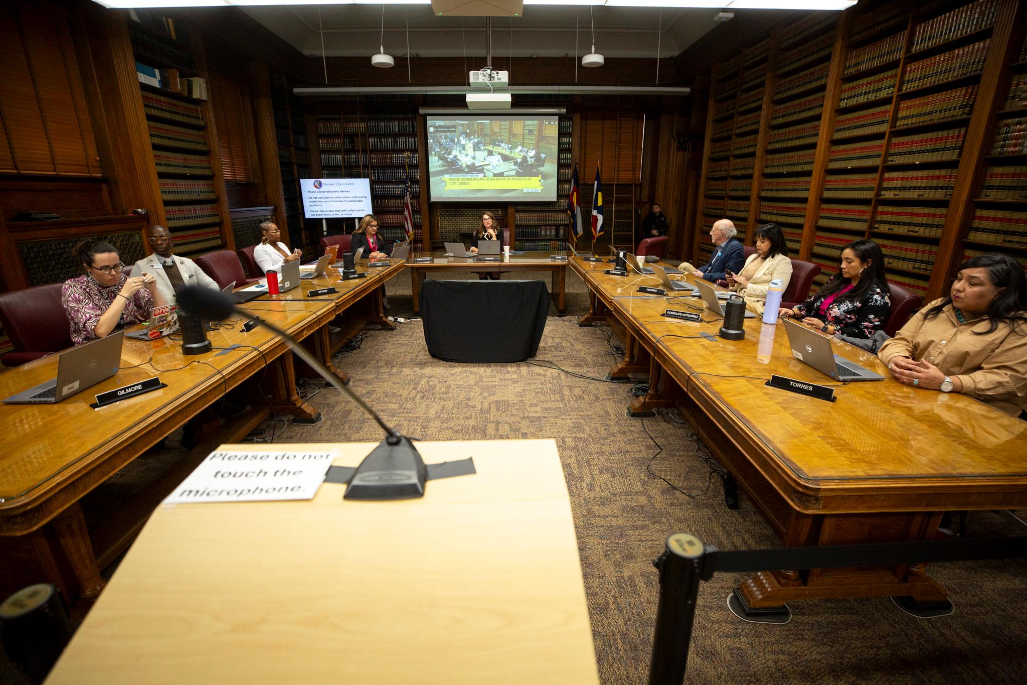 A view over a podium at a group of people in business attire sitting around wooden tables, in a warmly lit room lined with filled bookshelves.