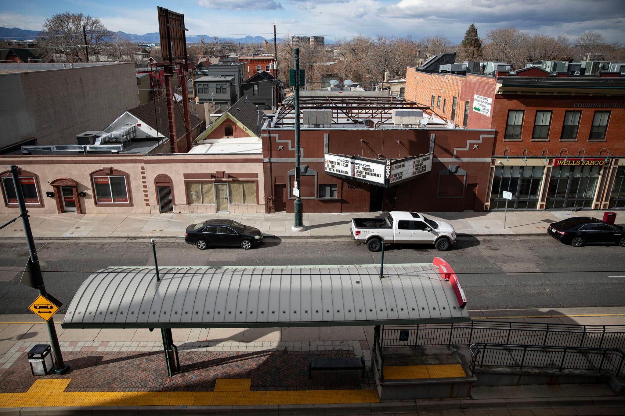 A view looking down at squat brick buildings and a train station along an asphalt street. A sign on the building to the right reads "ROXY" and features marquee listings.