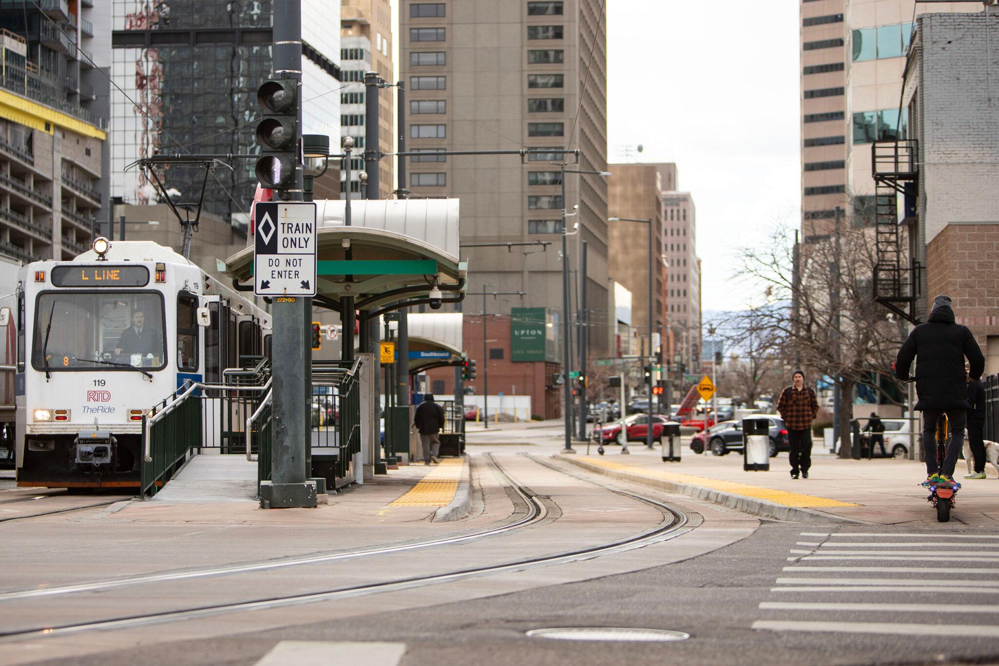 Train tracks curve into the frame and shoot towards the horizon. A train station, with a white train parked next to it, fills the left side of the frame as people walk, drive and scoot through the right. Tall buildings rise in the background.