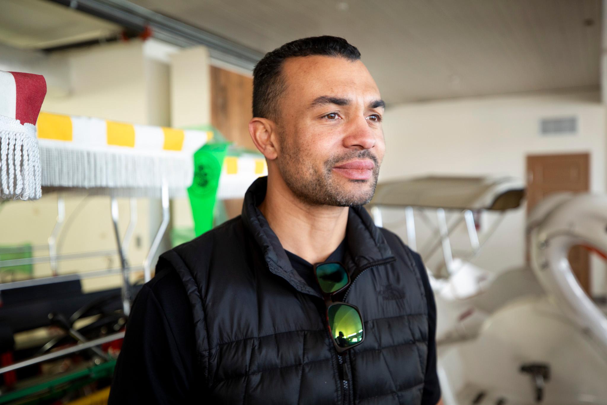 A man in a black jacket stands in a white room, looking emotional. The shapes of swan boats can be seen behind him.