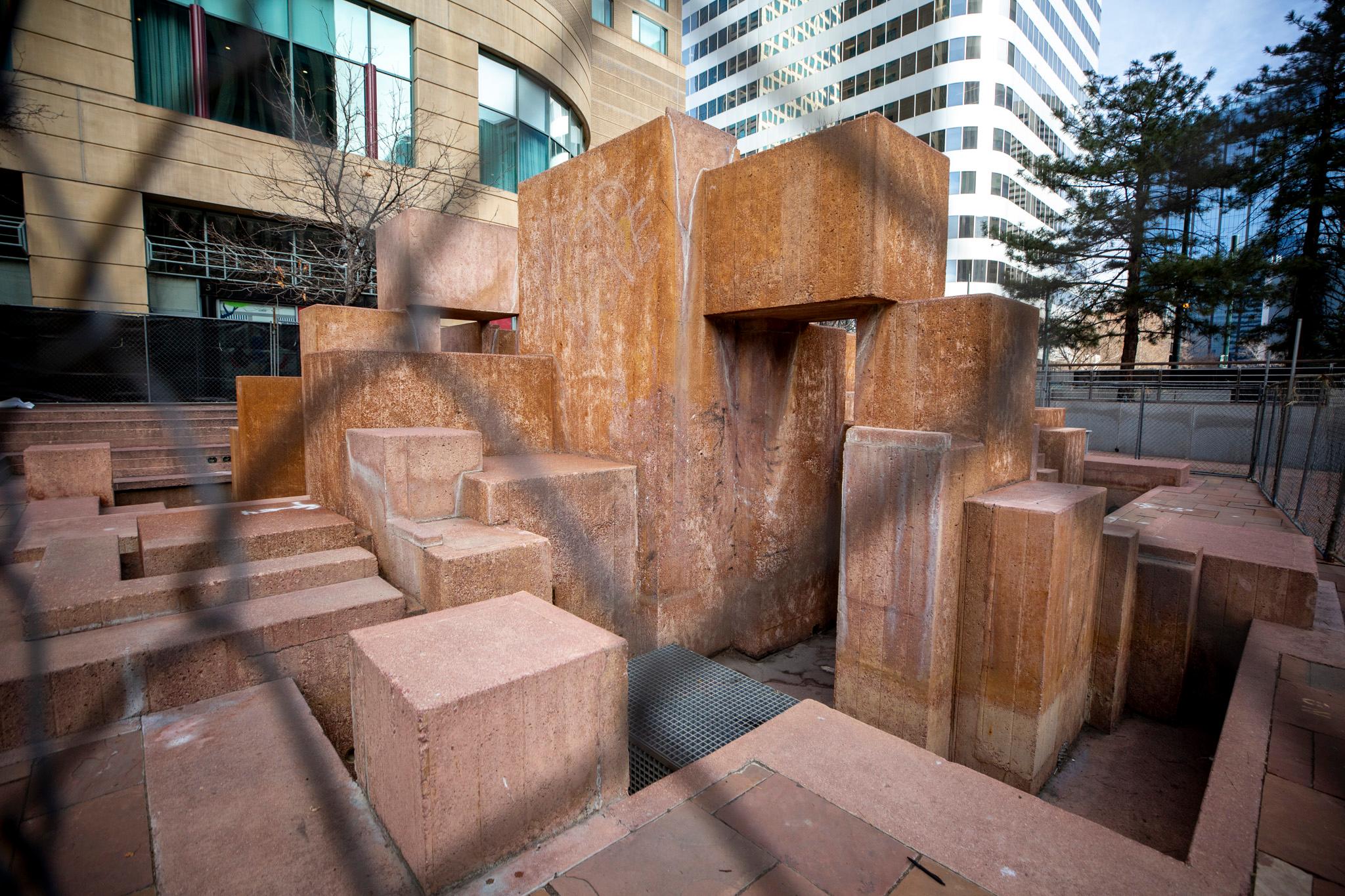 Cubes of stone are stacked in a plaza, seen through chain-link fencing.