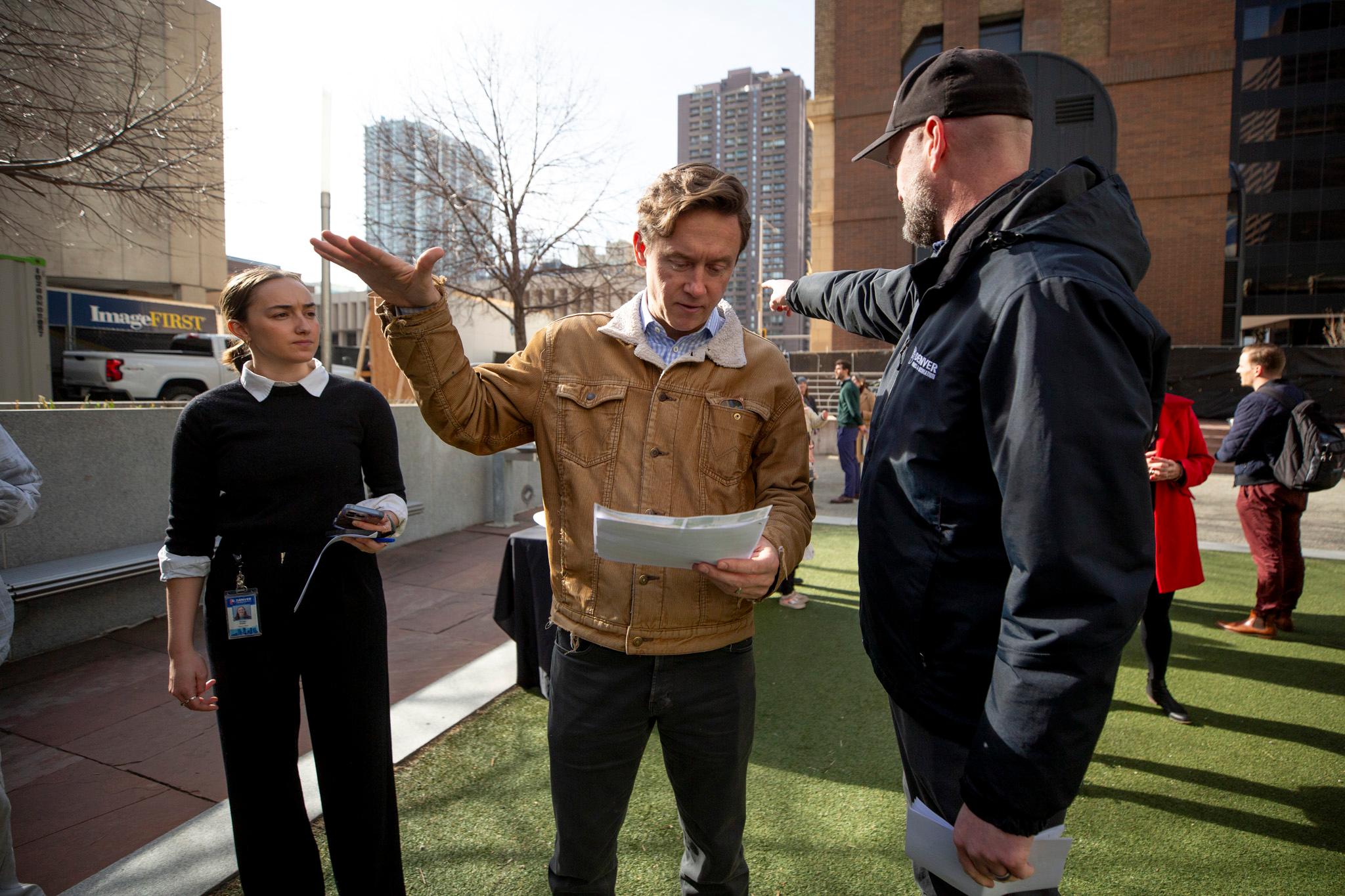 A man in a brown aviator jacket lifts a hand as he studies a piece of paper. Another man in a ball cap points behind him.