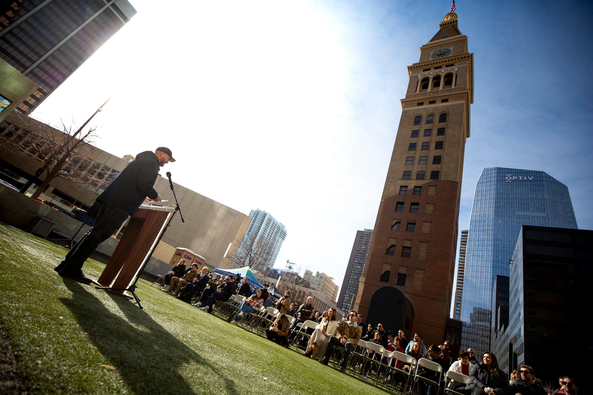 A man speaks at a podium in front of a crowd. A large clocktower rises far above them.