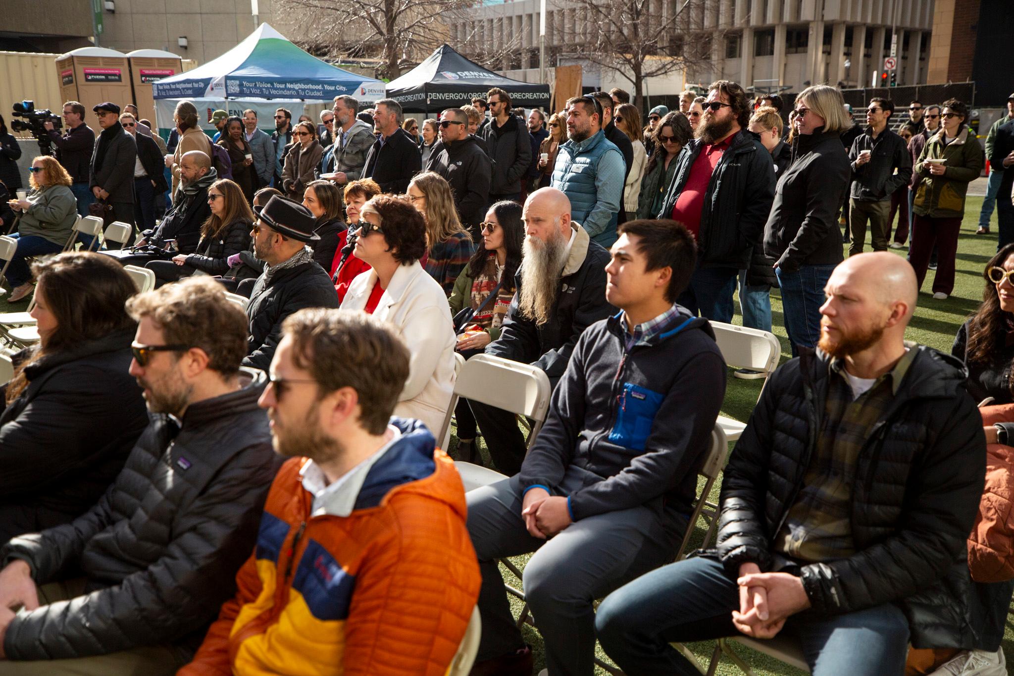 A crowd of people stand and sit outside, watching something out of frame.
