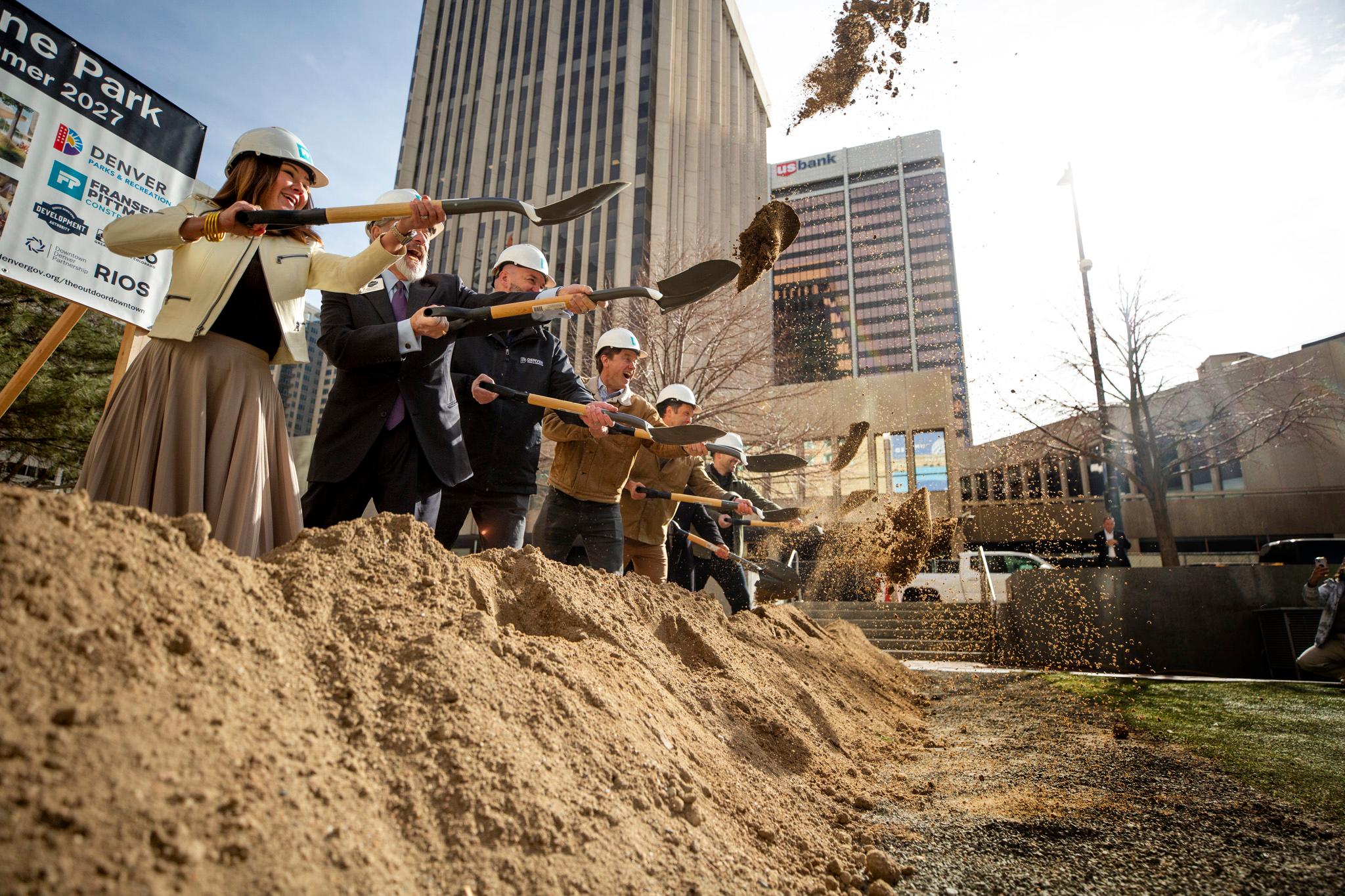 A group of people in hard hats fling shovels toward the sky as dirt rains down around them.