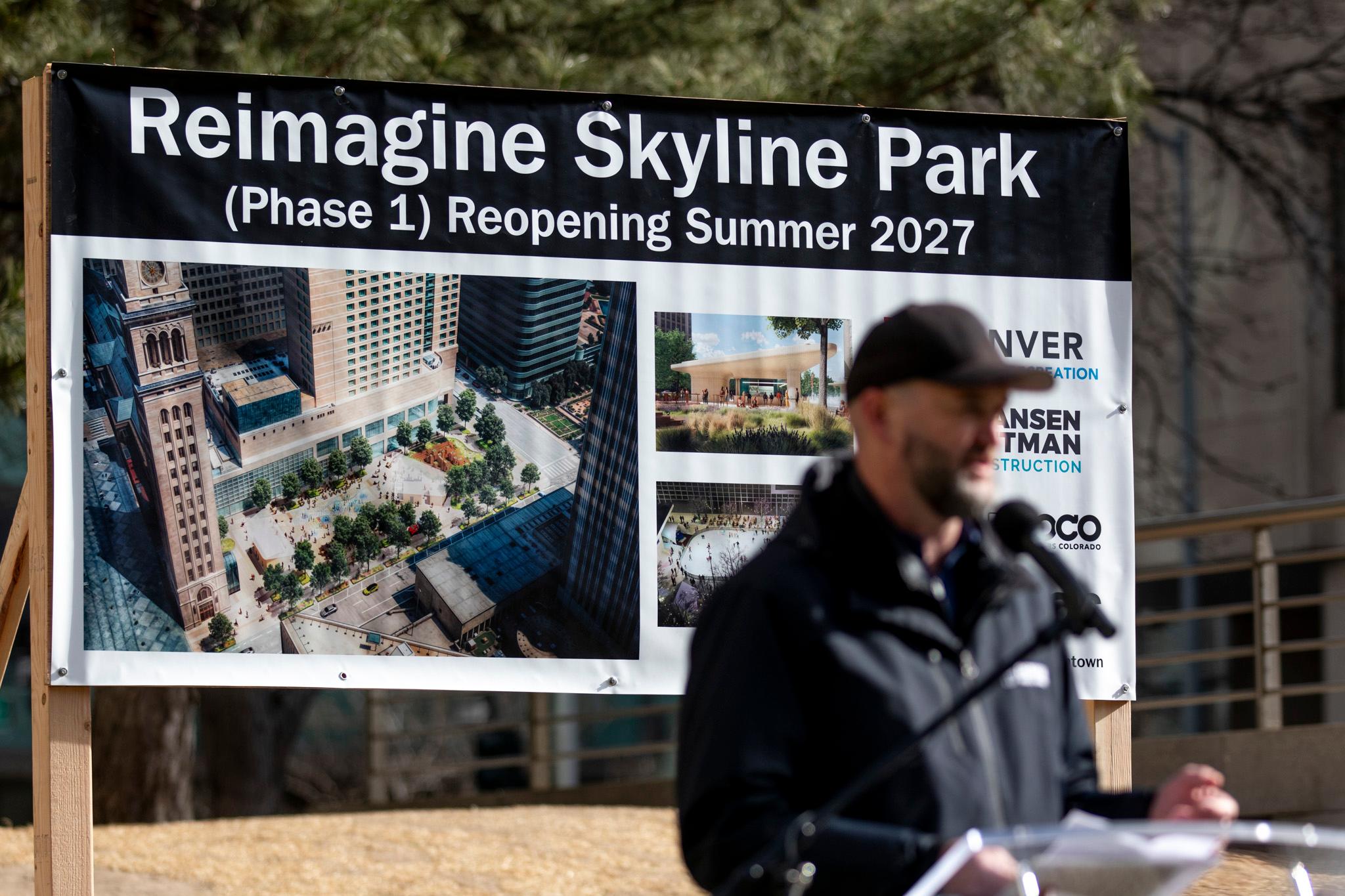 A man in a hat speaks at a podium. He's out of focus, and instead the image centers on a sign behind him that reads "Reimagine Skyline Park."