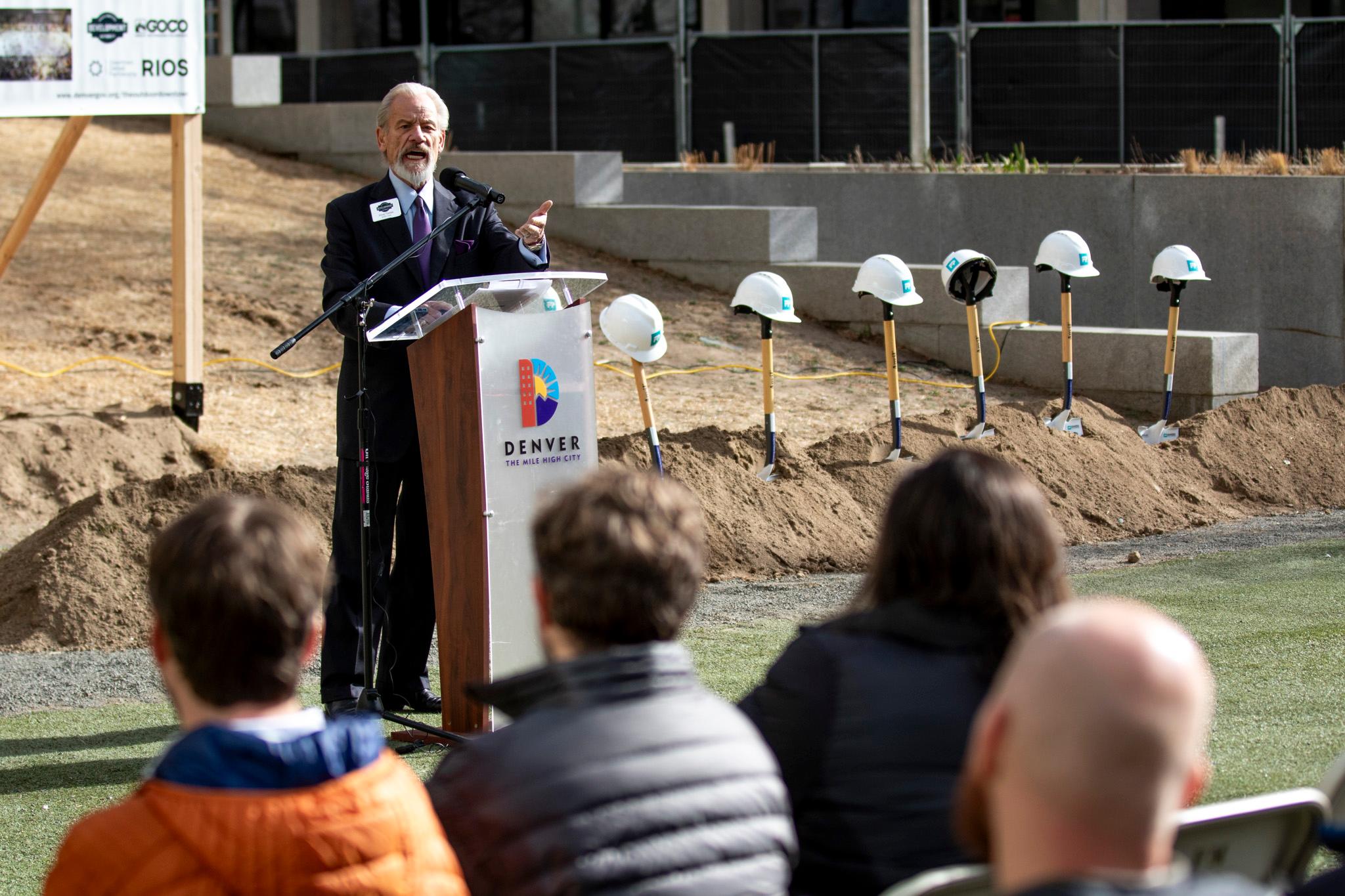 A man in a suit stands at a podium, speaking with a wide-open mouth as shovels sit in dirt behind him, each with a hard hat crowning its handle.