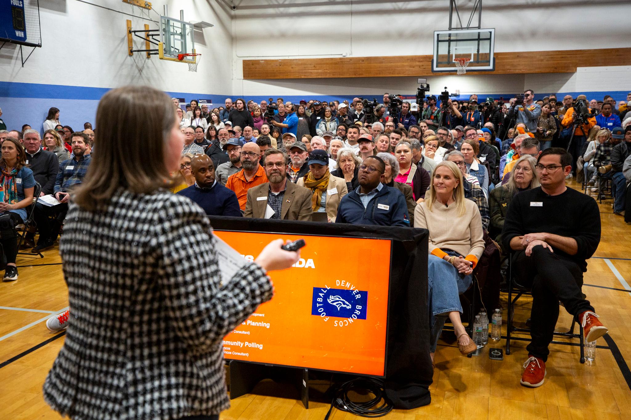 A gym is filled with people, watching a woman in the foreground, out of focus, as she speaks with her hands.