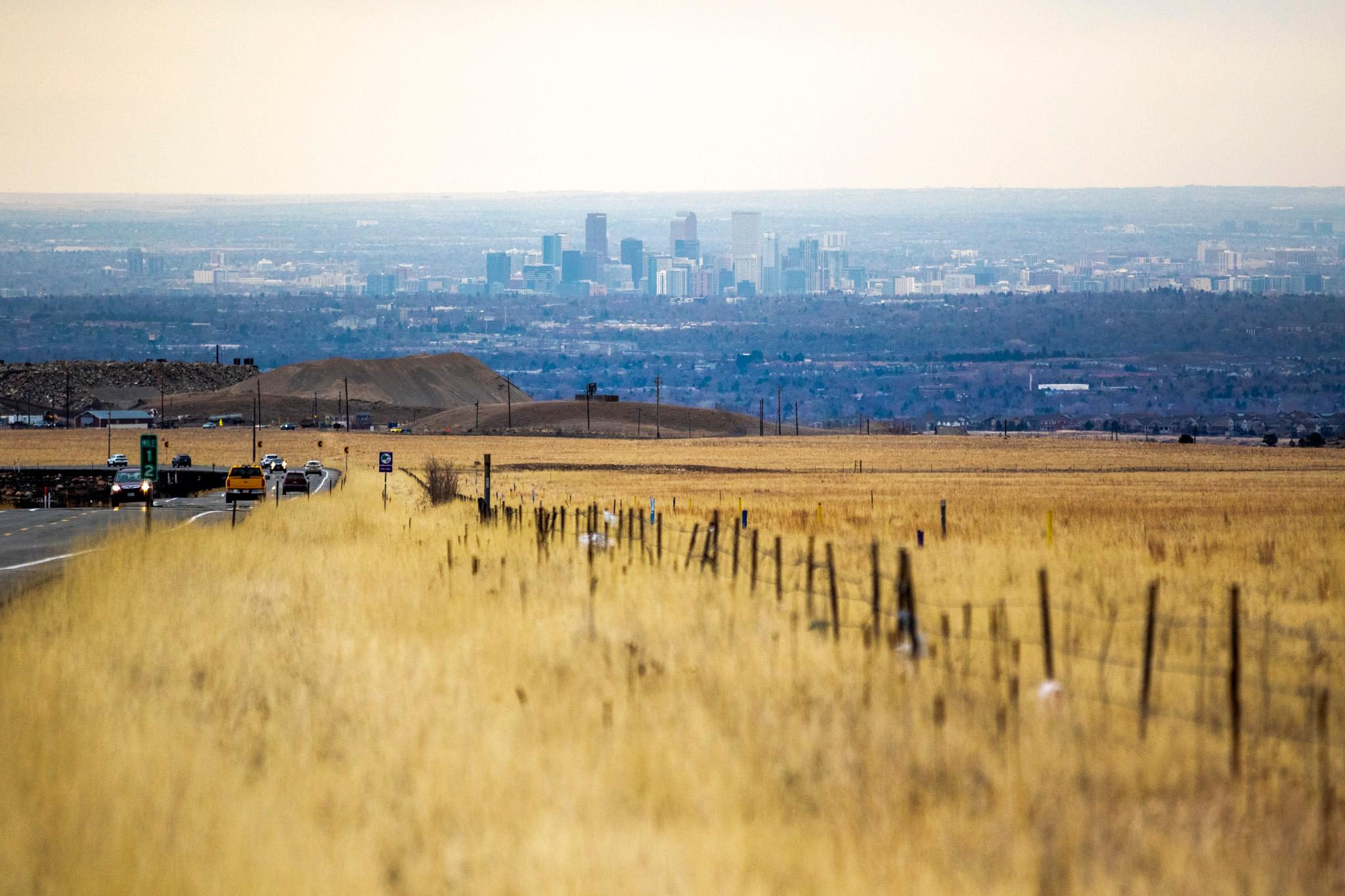 A city stands above a flat plain, seen from far away. The foreground is filled with yellow wheat.