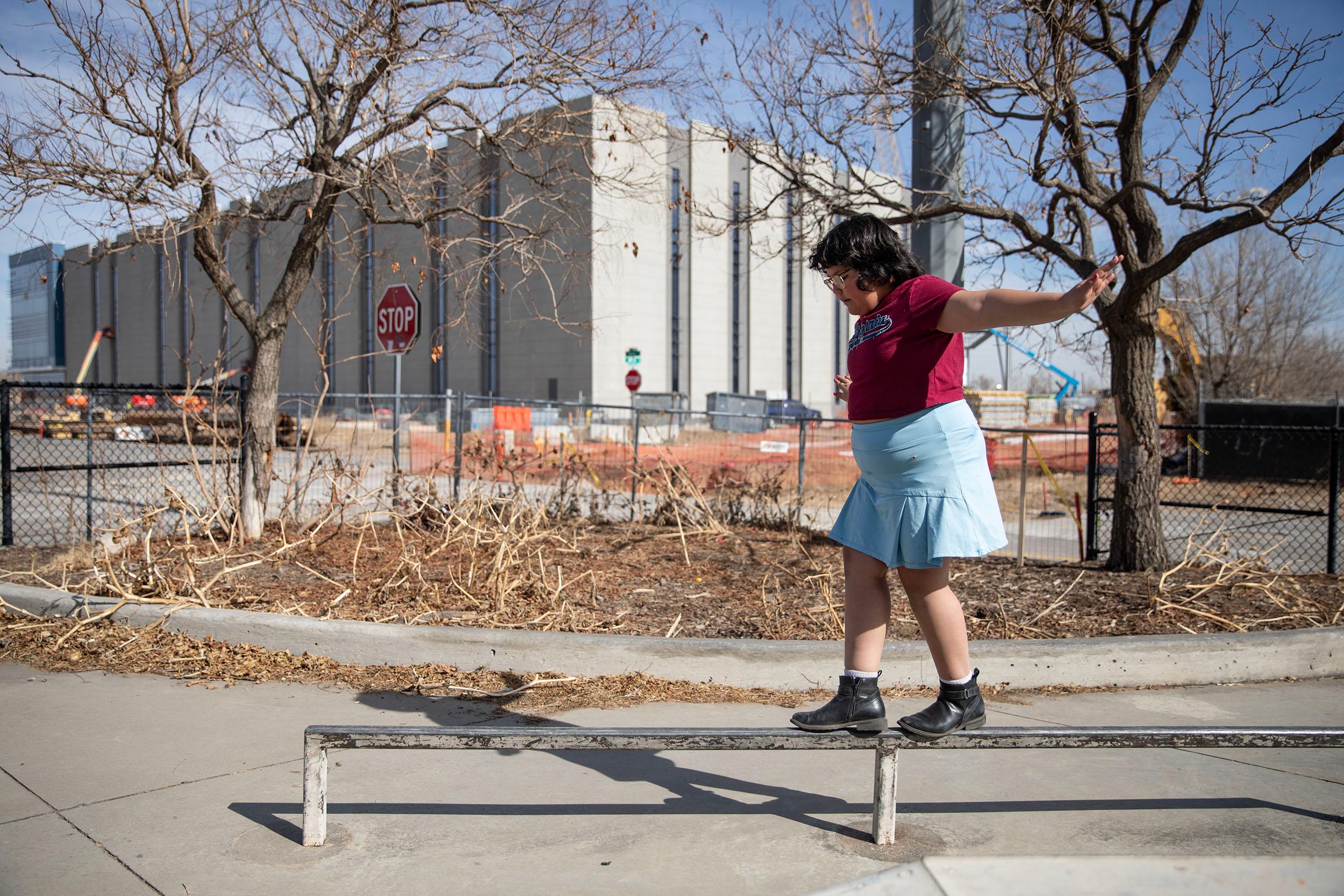 A girl in a blue skirt tiptoes over a rail on a sunny day.