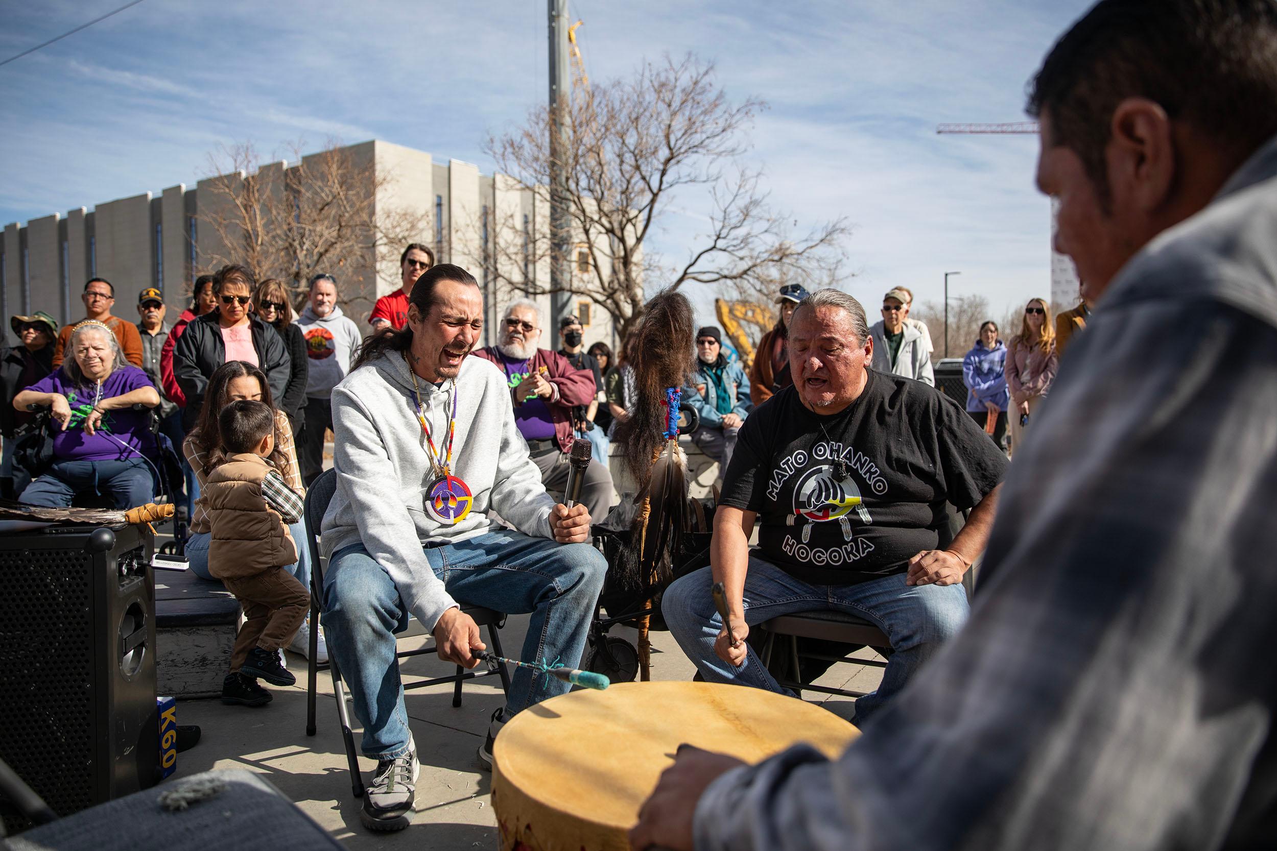 Men sit around a round drum, mouths open as they sing, with a crowd surrounding them.