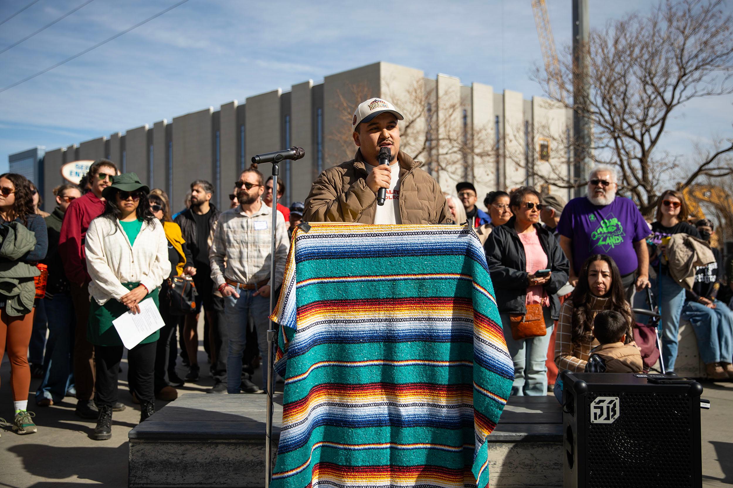 A man in a cap speaks at a microphone, at a podium covered by a yellow and blue textile.