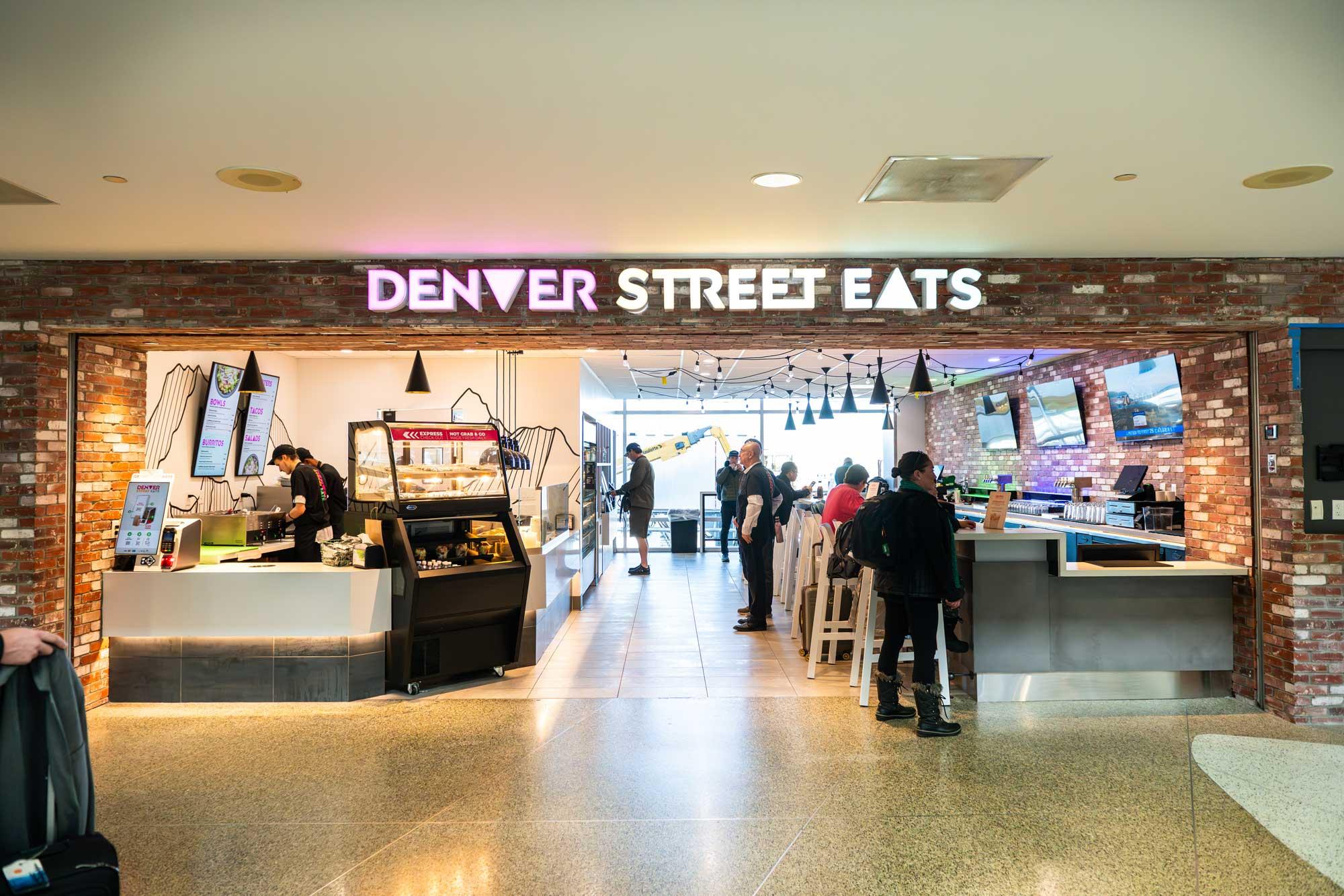 A brick facade with a sign reading "Denver Street Eats" opens into an airport restaurant with a food counter on the left and a bar on the right.