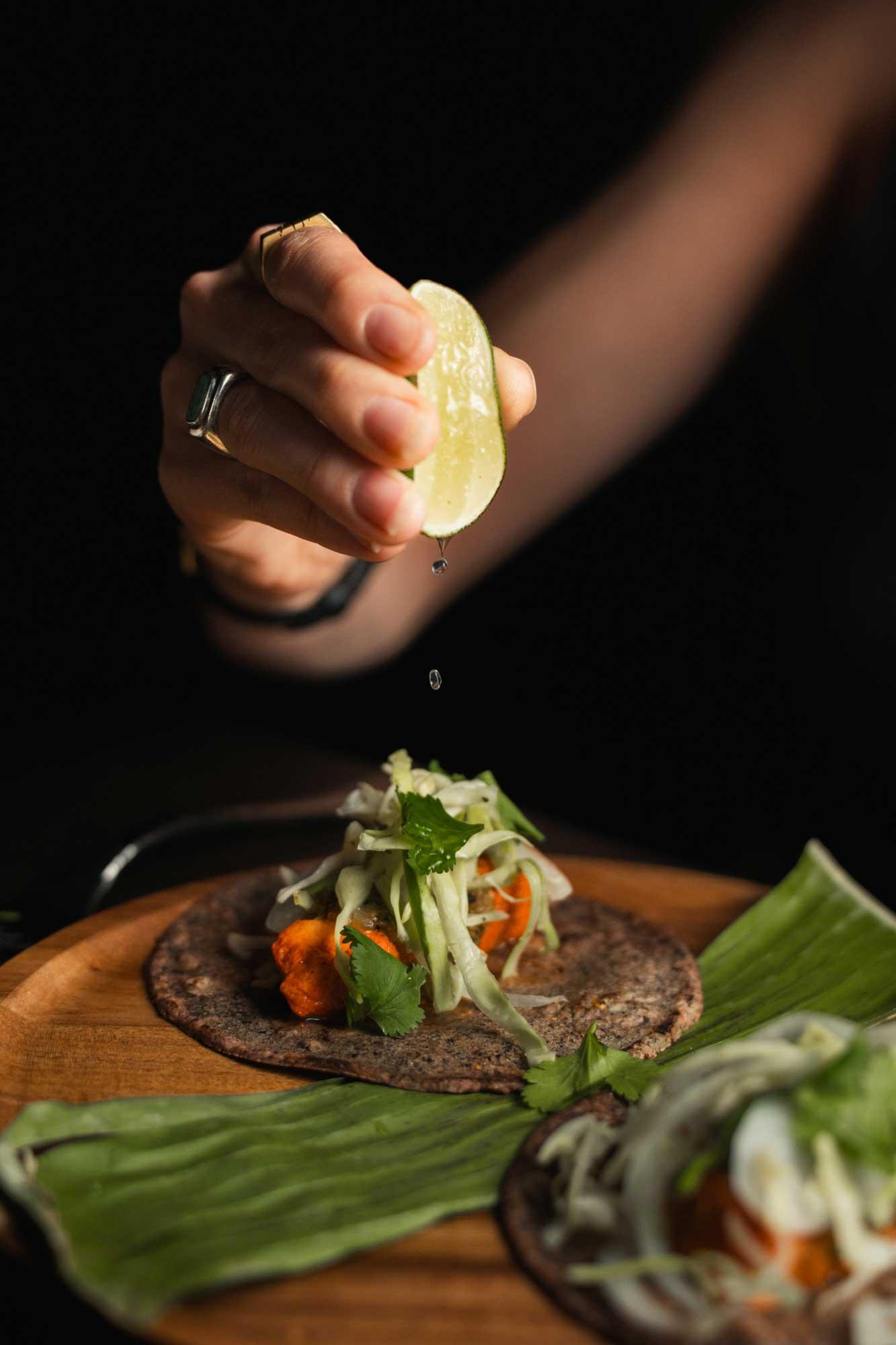 A hand squeezes lime over two tacos on a warm brown plate. The tacos are stacked with cabbage and other vegetables on blue corn tortillas.