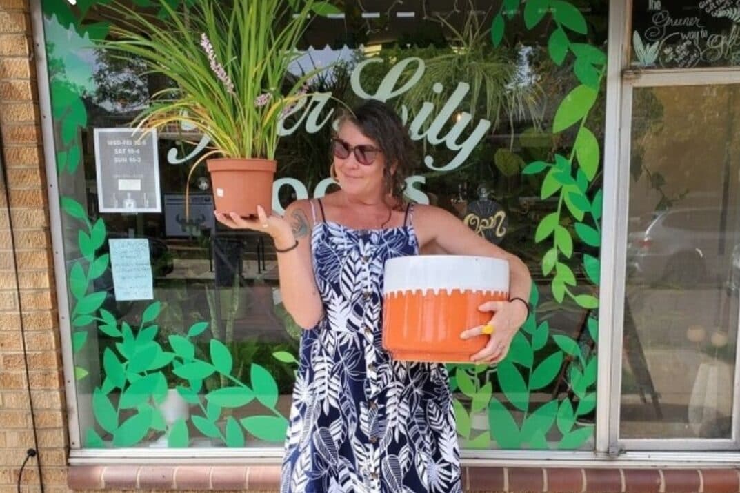 A woman in a blue-and-white print dress stands outside a window painted with green leaves. She holds a large pot in each hand.