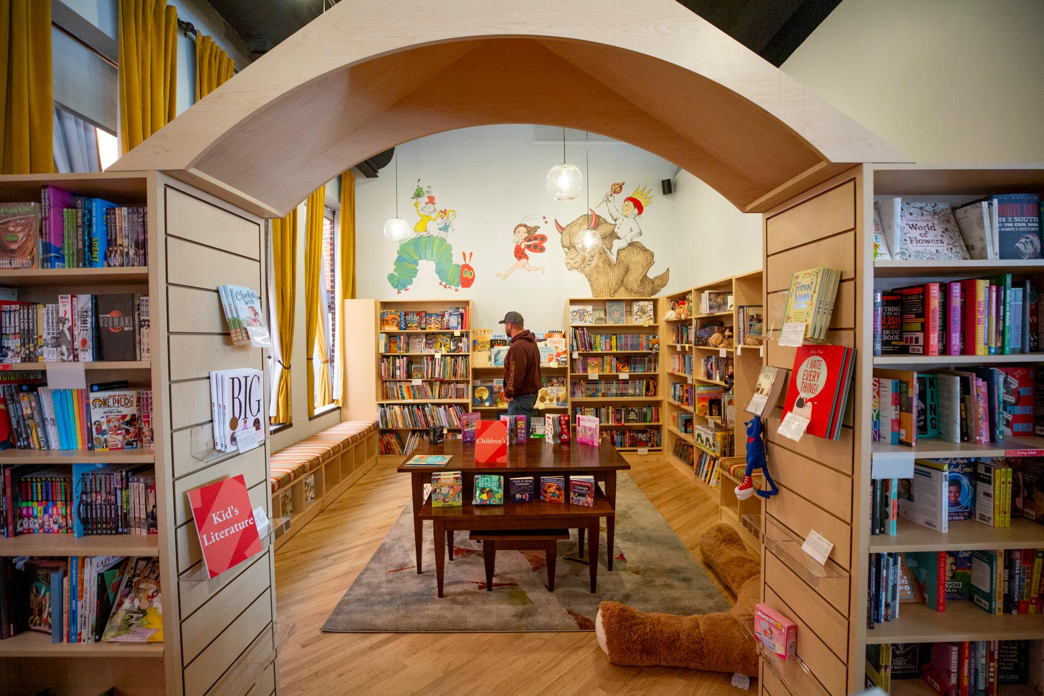 A man in a hat stands in a well lit room lined with filled bookshelves. Cartoon characters are drawn on the wall above.