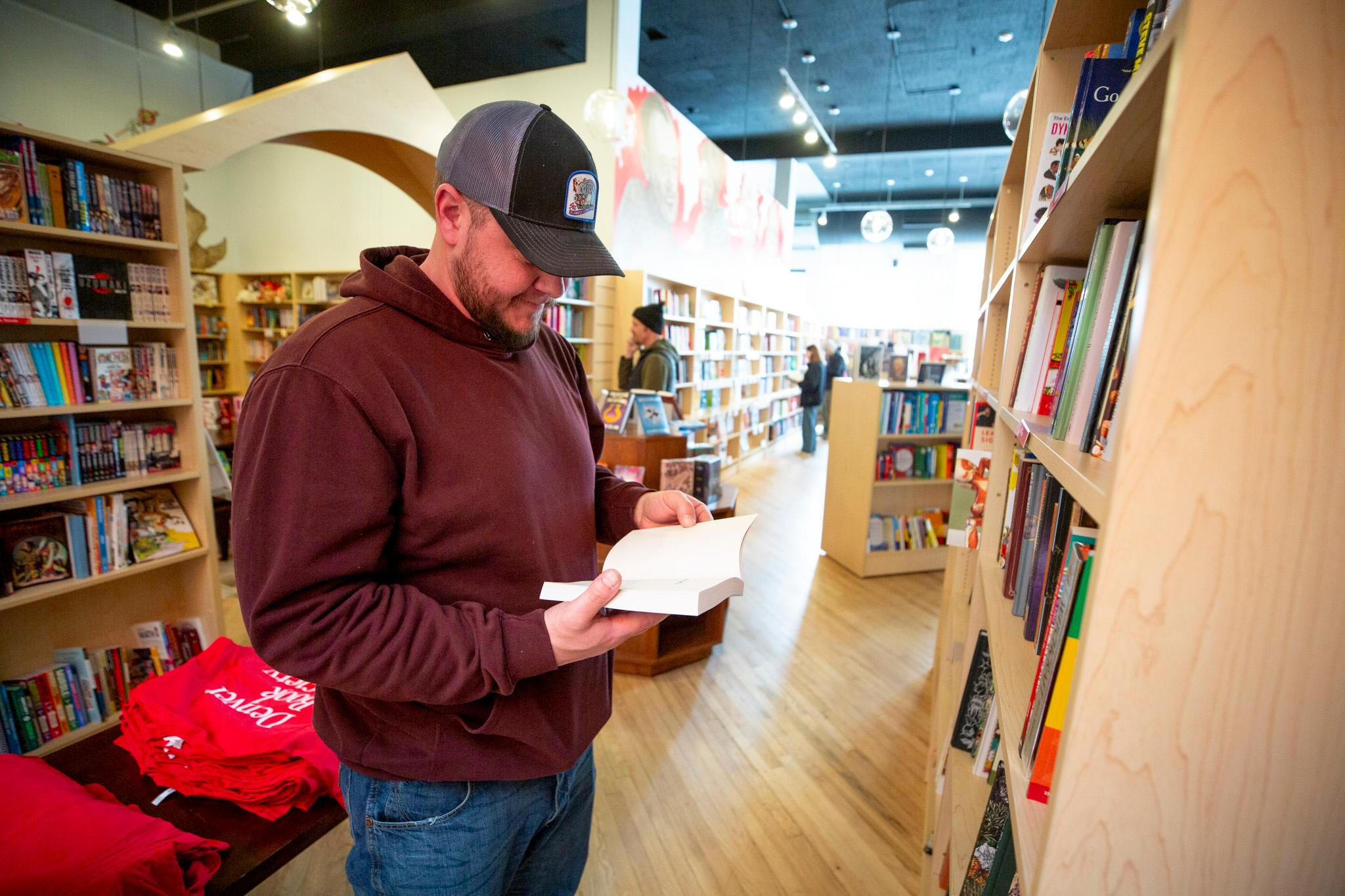 A man peers into an open book in a room filled with wooden bookshelves and colorful covers.