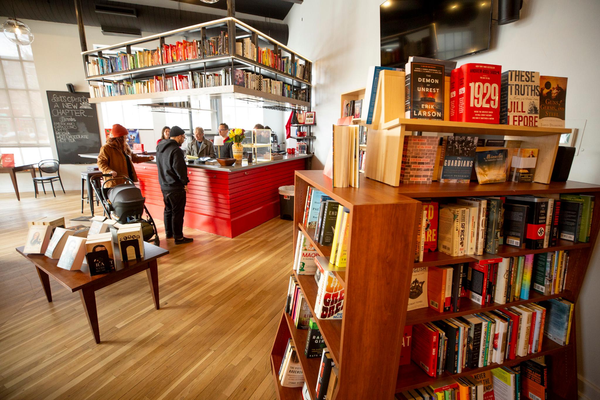 People stand at a cafe counter in awarmly lit room. A full bookshelf fills the right side of the frame.