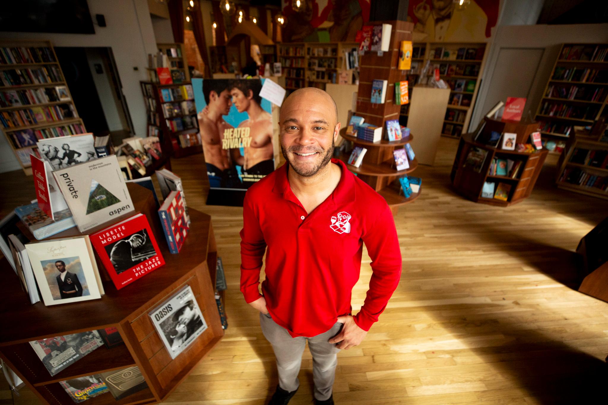 A man in a red shirt smiles at the camera, in a warmly lit room of wooden floors and bookshelves filled with colorful tomes.