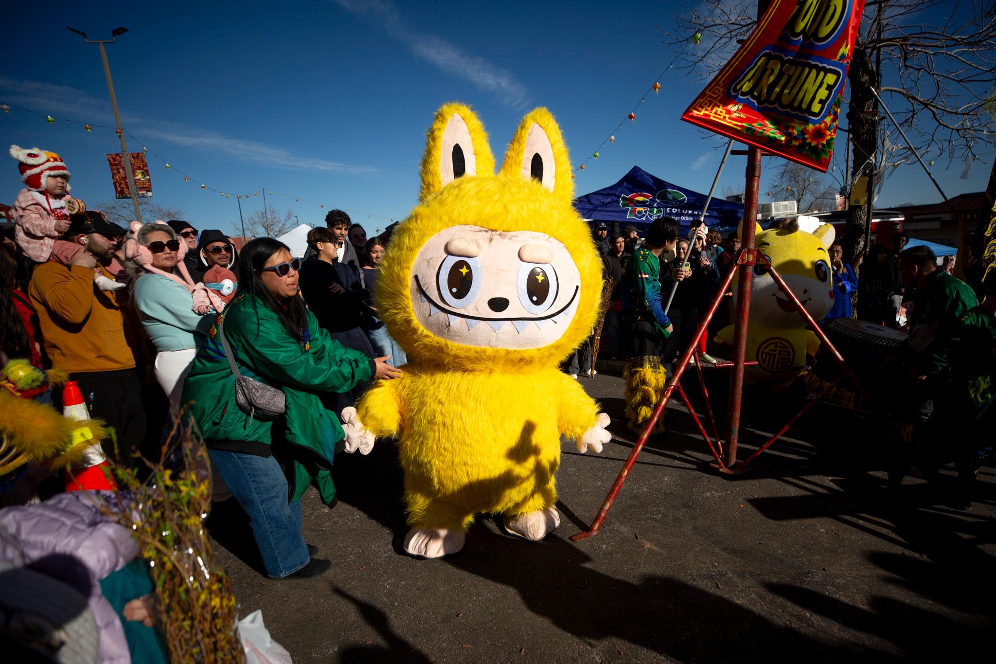 A human-sized Lububu — a cartoon-eyed gremlin with bunny ears, sharp teeth and a bright yellow coat of fur — stands in a crowd of people.