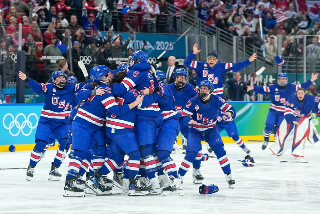 At least a dozen hockey players in blue-and-red uniforms embrace on ice, with several teammates skating over to meet the group. The crowd behind them reacts in a mix of excitement and sadness.