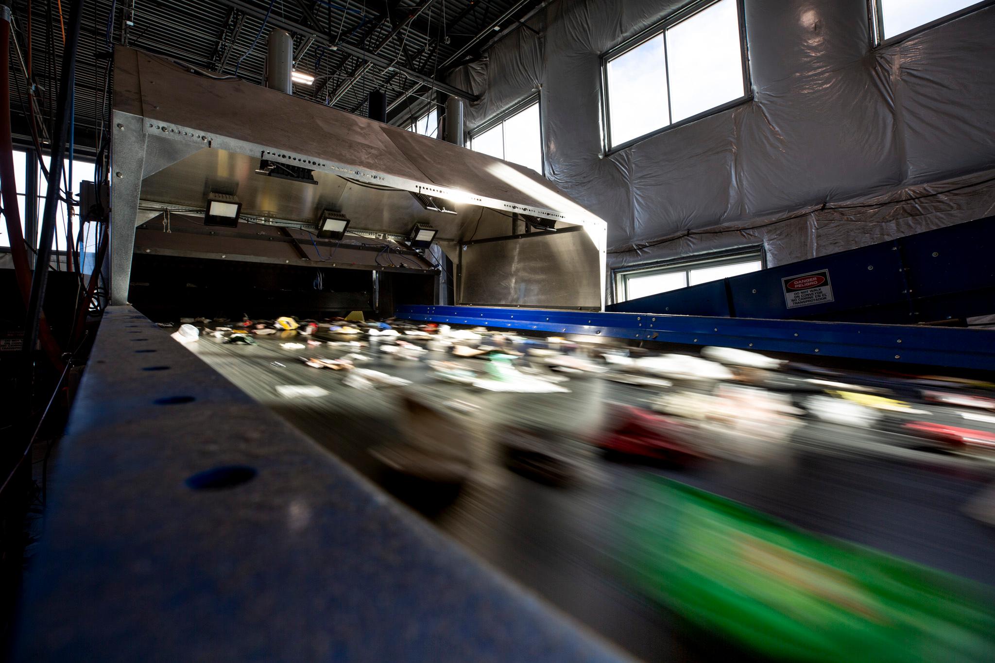 A low view of a conveyor belt. Trash atop it blurs through across the frame. It's all heading towards a black abyss at the end of the line.