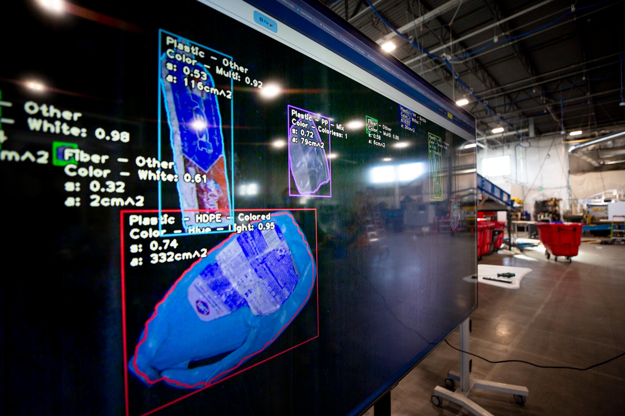 A giant screen in a warehouse shows plastic trash — a blue bottle and a toothpaste tube — which are annotated with computer generated squares and text.