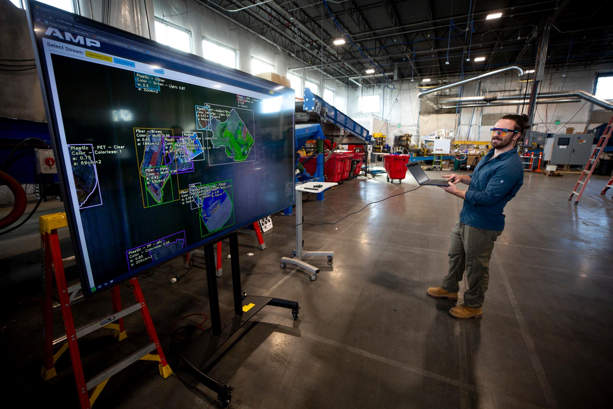 A man in safety glasses watches an enormous screen in a concrete-floored warehouse.